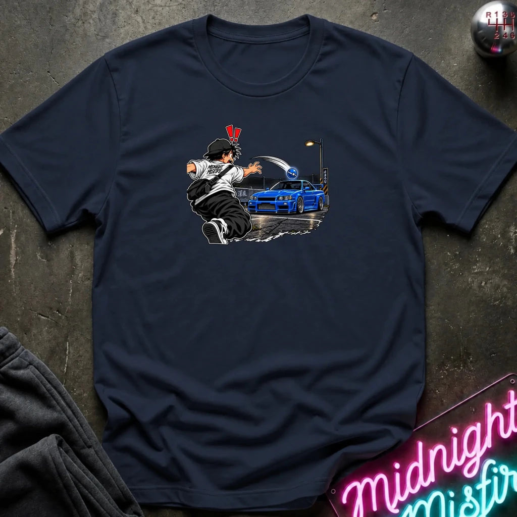 A navy blue short-sleeve t-shirt with a graphic print of a street artist spray-painting a blue sports car, displayed on a dark textured background with a neon sign and sweatpants.
