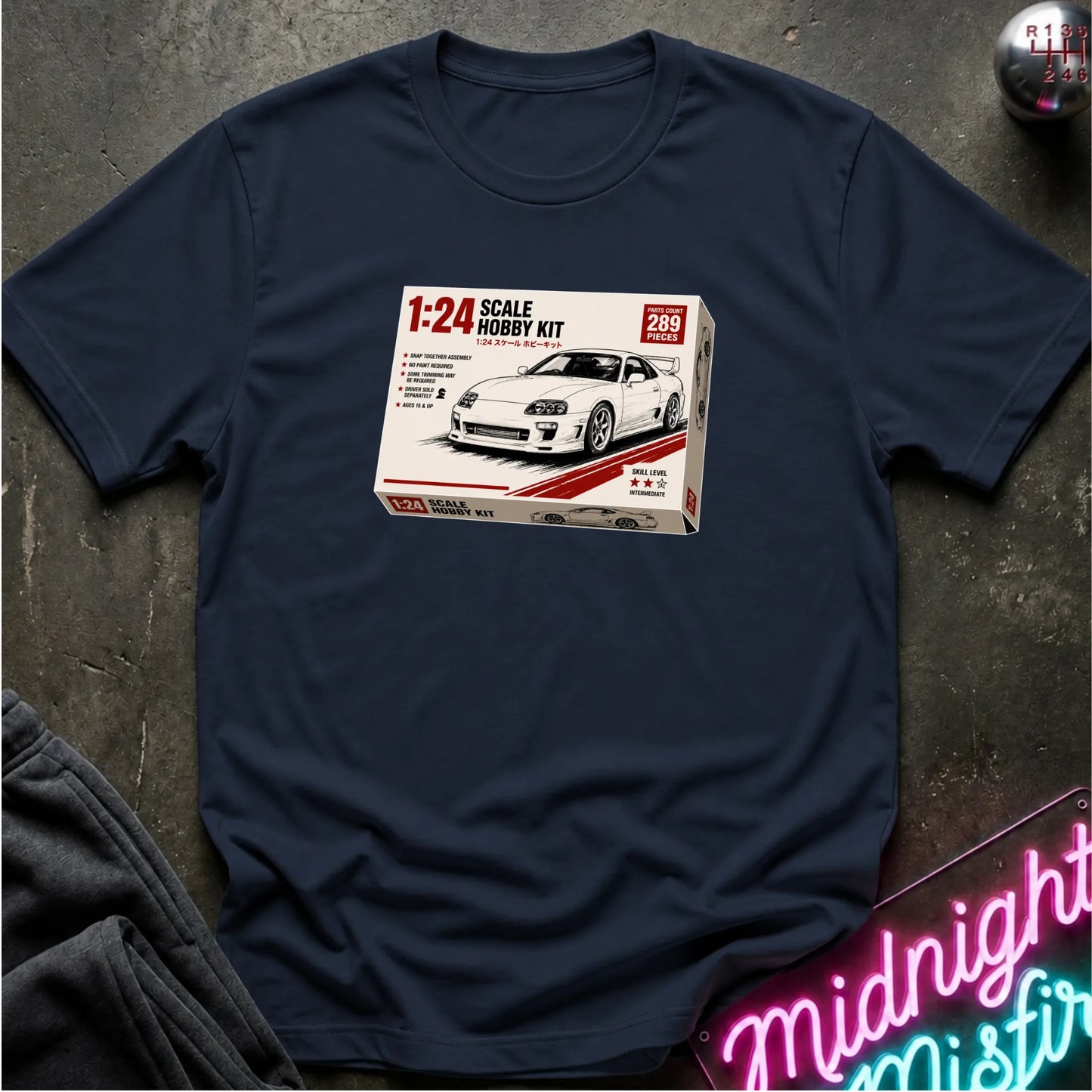 A navy blue short-sleeve t-shirt laid flat on a dark textured surface, featuring a printed graphic of a 1:24 scale model kit box for a white sports car. A neon sign is partially visible in the bottom right corner.