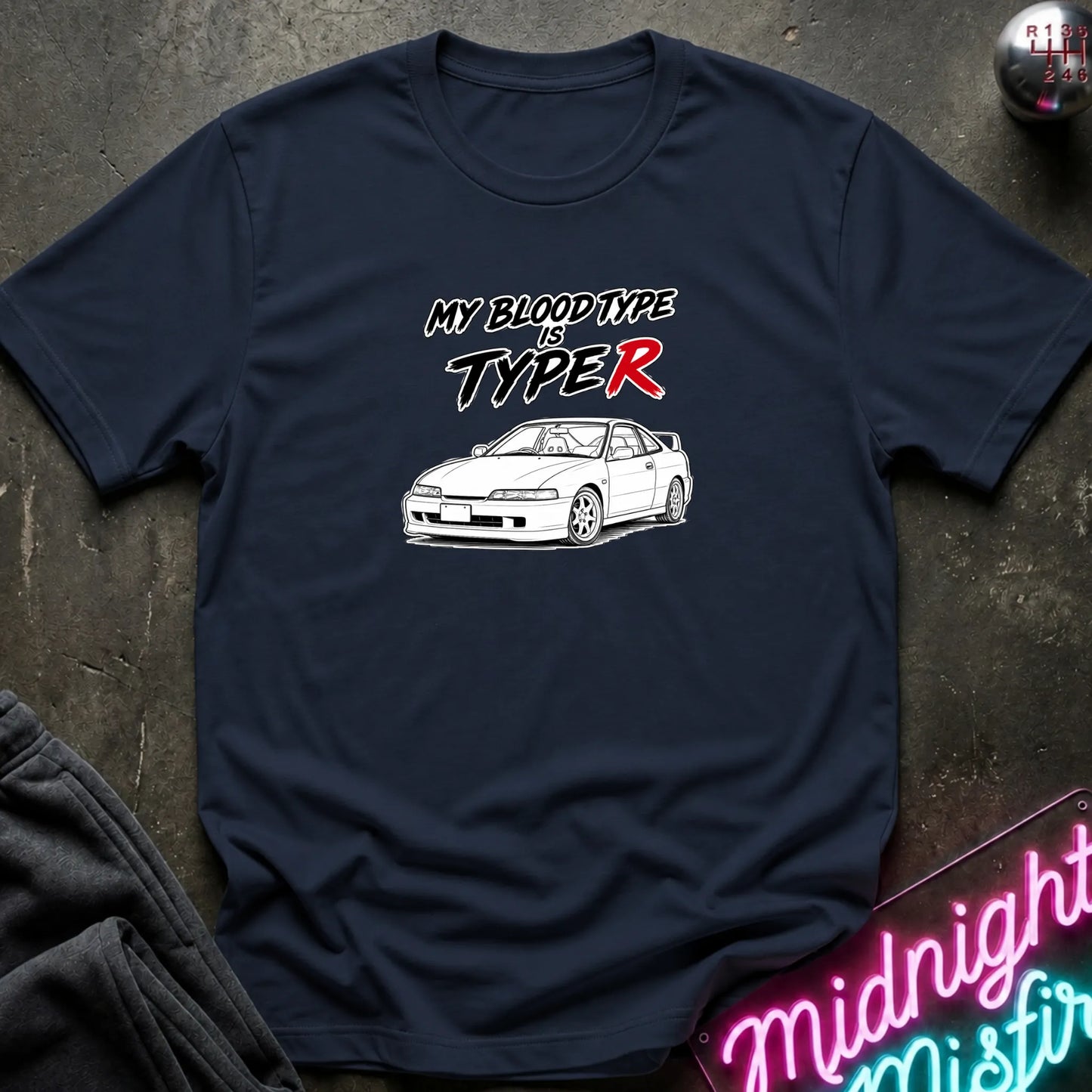 A navy blue graphic t-shirt laid flat on a dark textured surface. The shirt features a white line art drawing of a car and the text 'MY BLOODTYPE IS TYPER' in stylized white and red lettering.