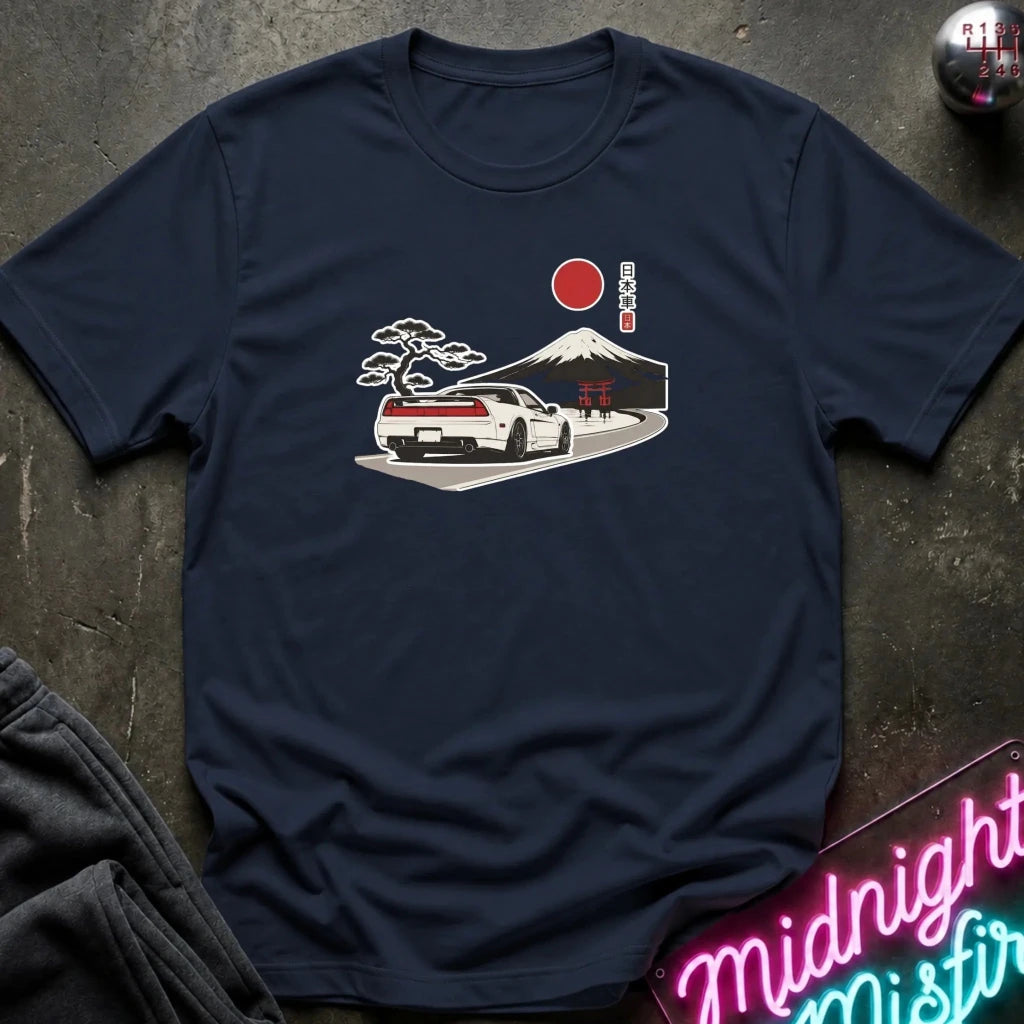 A navy blue short-sleeve t-shirt laid flat on a dark textured surface, featuring a graphic print of a white sports car on a road with a red sun, Mount Fuji, and a pine tree in a Japanese-style illustration.