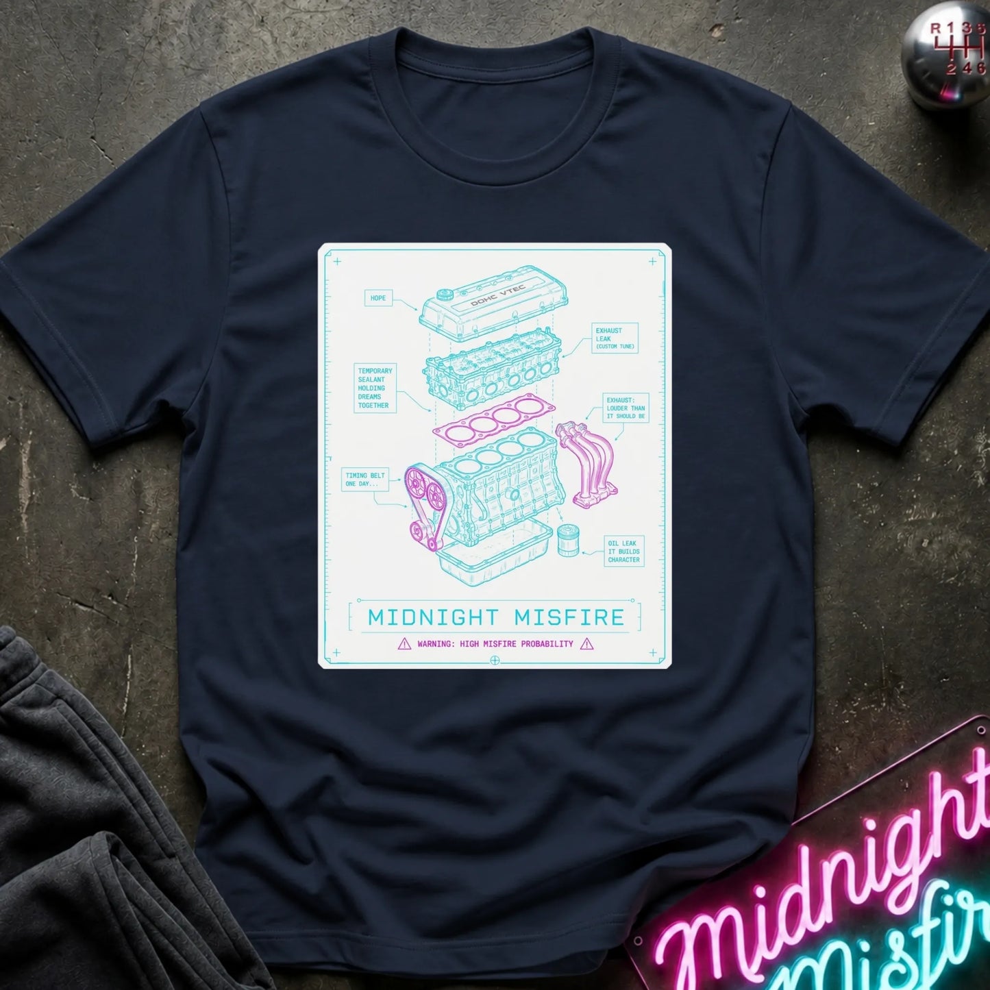 A navy blue short-sleeve graphic T-shirt laid flat on a dark textured surface. The shirt features a large rectangular print with a light blue engine diagram and the text 'MIDNIGHT MISFIRE.