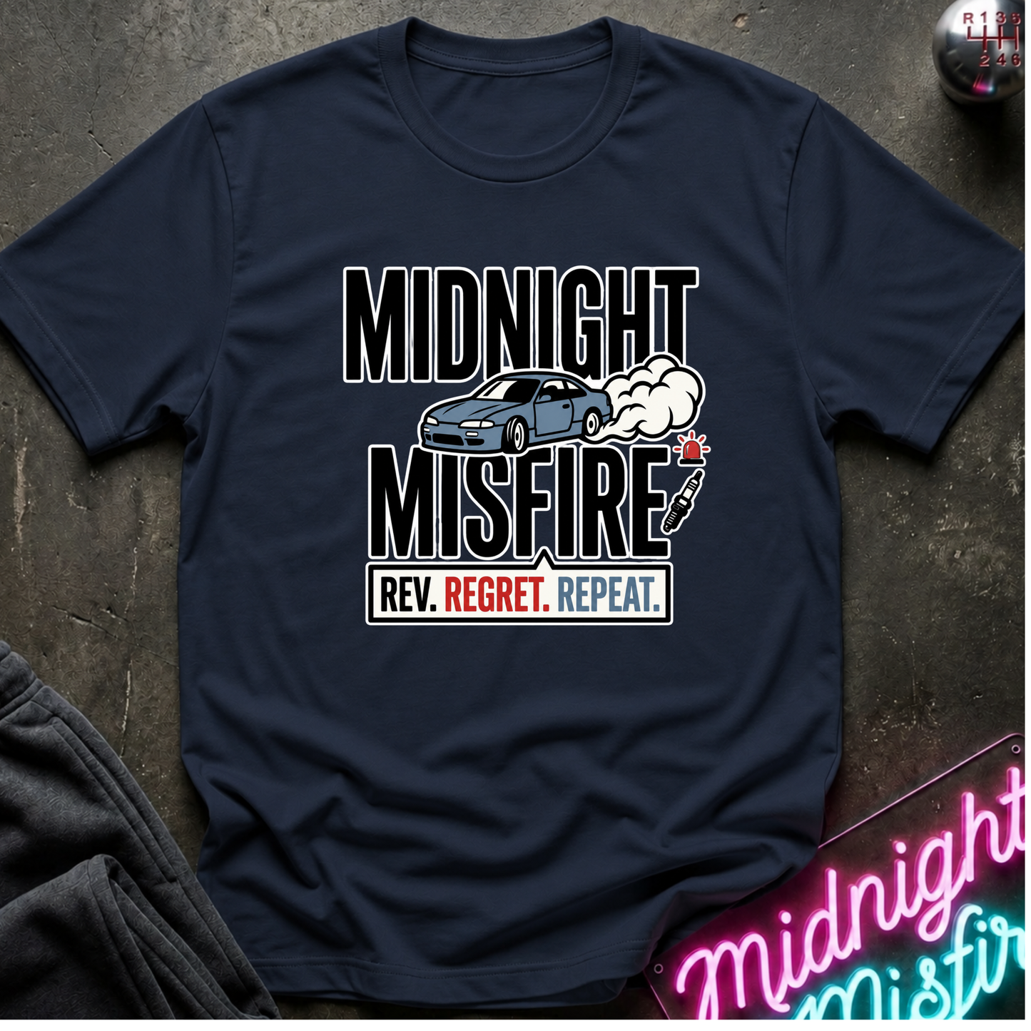 A navy blue crewneck t-shirt laid flat on a dark textured surface. The shirt features a graphic print with the words 'MIDNIGHT MISFIRE' in bold white letters, a blue sports car and smoke.