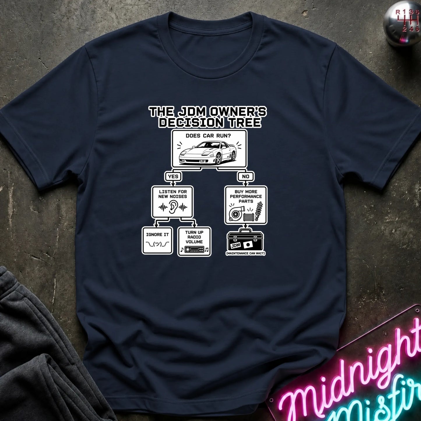 A navy blue short-sleeve graphic T-shirt laid flat on a dark textured surface. The shirt features a black printed decision tree titled 'THE JDM OWNER'S DECISION TREE' with illustrations and text related to car care and maintenance.