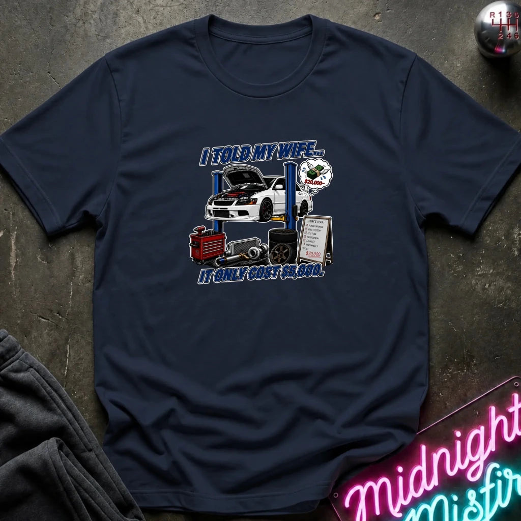 A navy blue graphic t-shirt laid flat on a dark textured surface. The graphic features a white sports car on a blue lift rack with the text 'I TOLD MY WIFE.