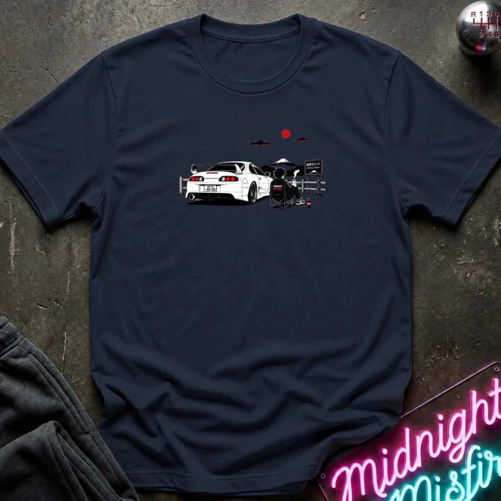 A navy blue short-sleeve graphic T-shirt laid flat on a dark textured surface. The shirt features a printed illustration of a white sports car with a red sun and mountain graphic above it.