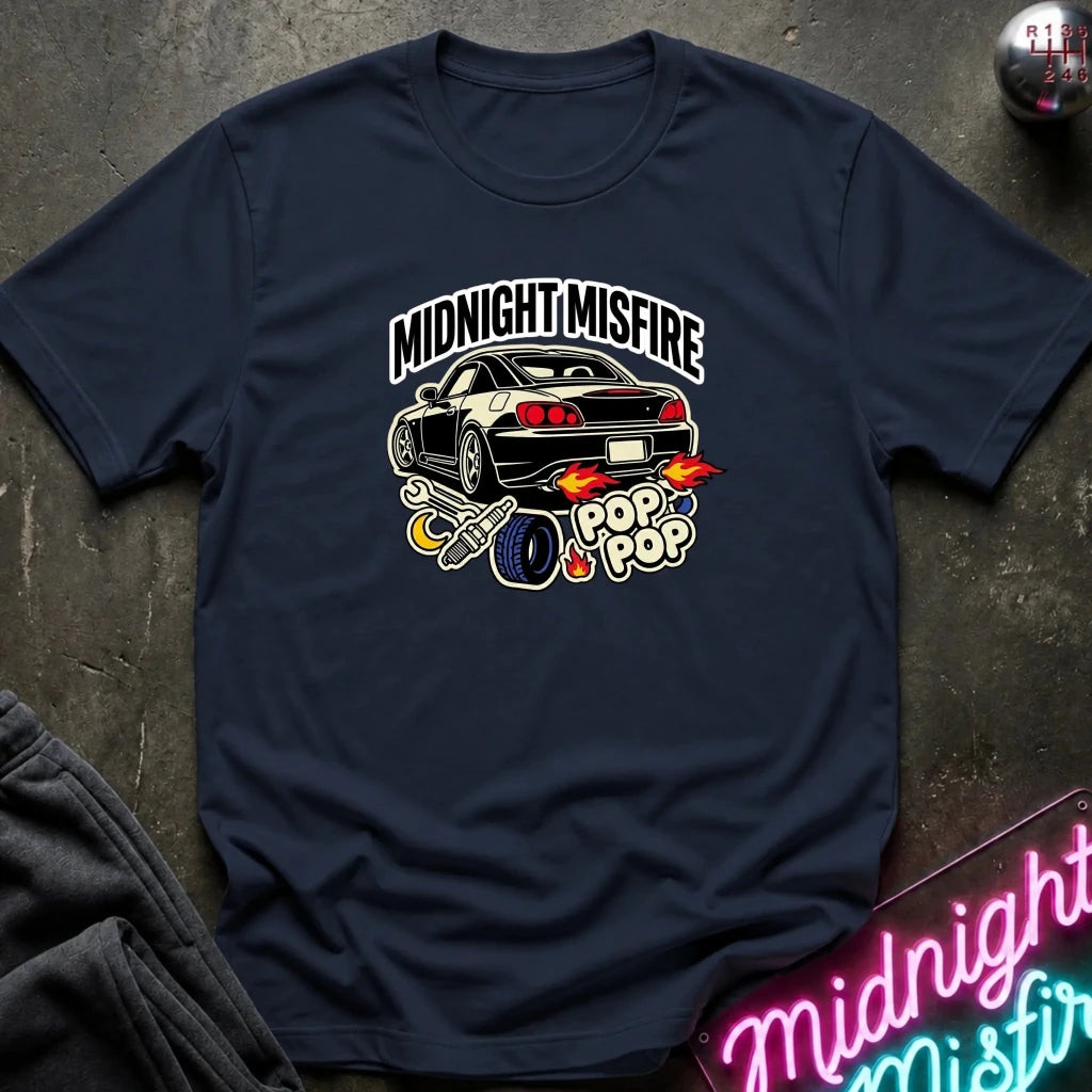 A navy blue graphic t-shirt laid flat on a dark textured surface. The shirt features a graphic of a black sports car with flames, a exhaust pipe, and the text 'MIDNIGHT MISFIRE' and 'POP POP'.