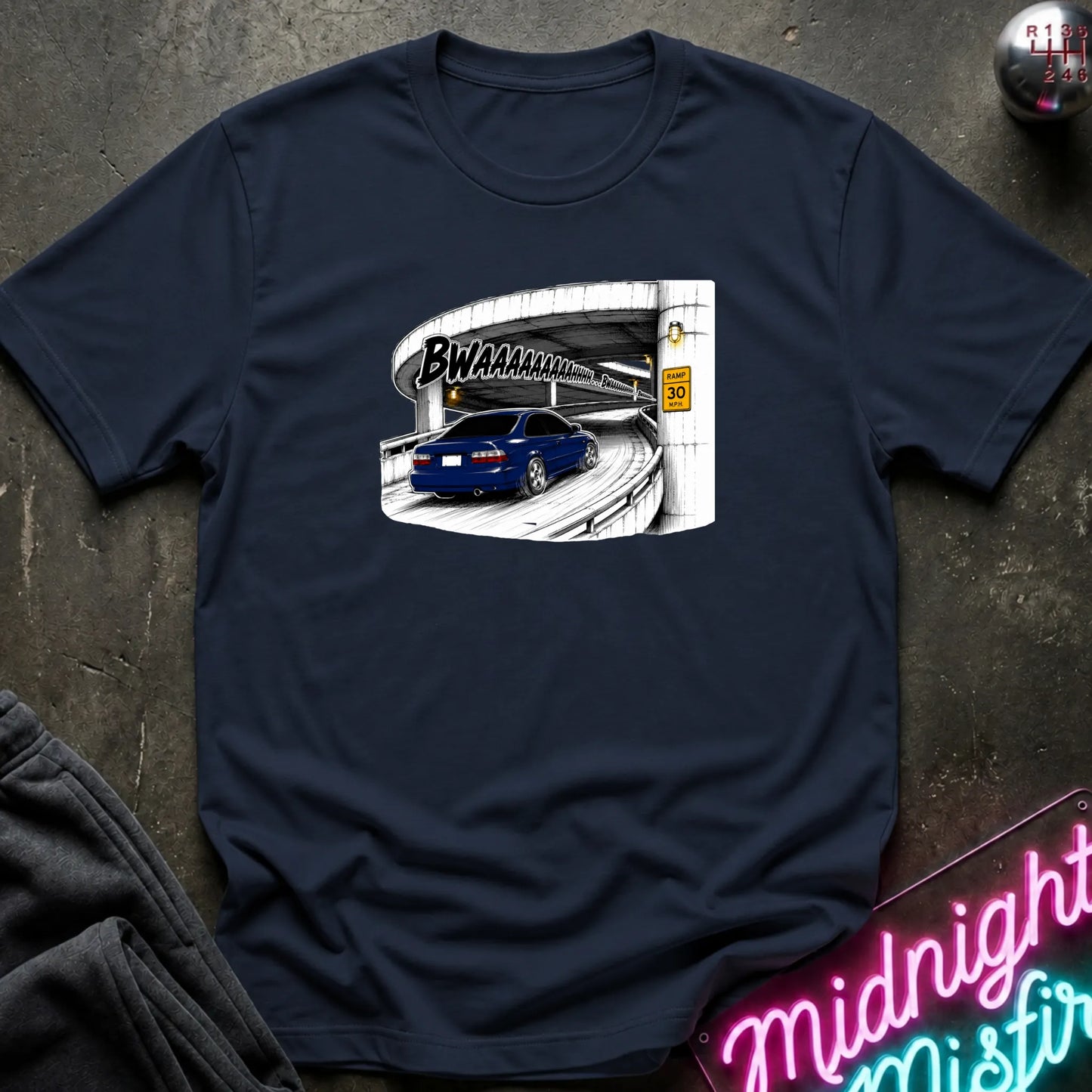 A navy blue short-sleeve crewneck t-shirt laid flat on a dark textured surface. The shirt features a graphic print of a blue car driving under a highway overpass with the text 'BWAHAHA'.