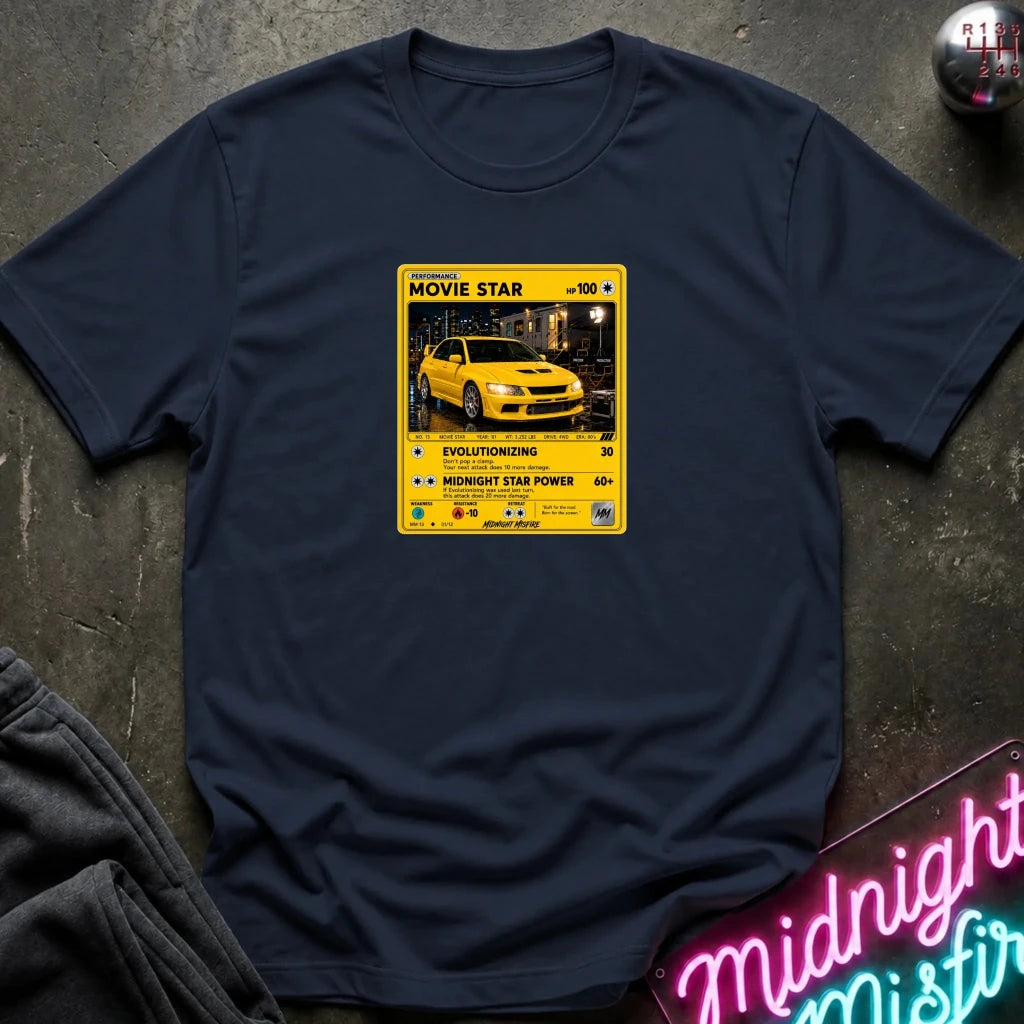 A navy blue short-sleeve t-shirt laid flat on a dark textured surface, featuring a yellow and black retro-style graphic on the chest with a yellow car photo and text including 'MOVIE STAR'. 