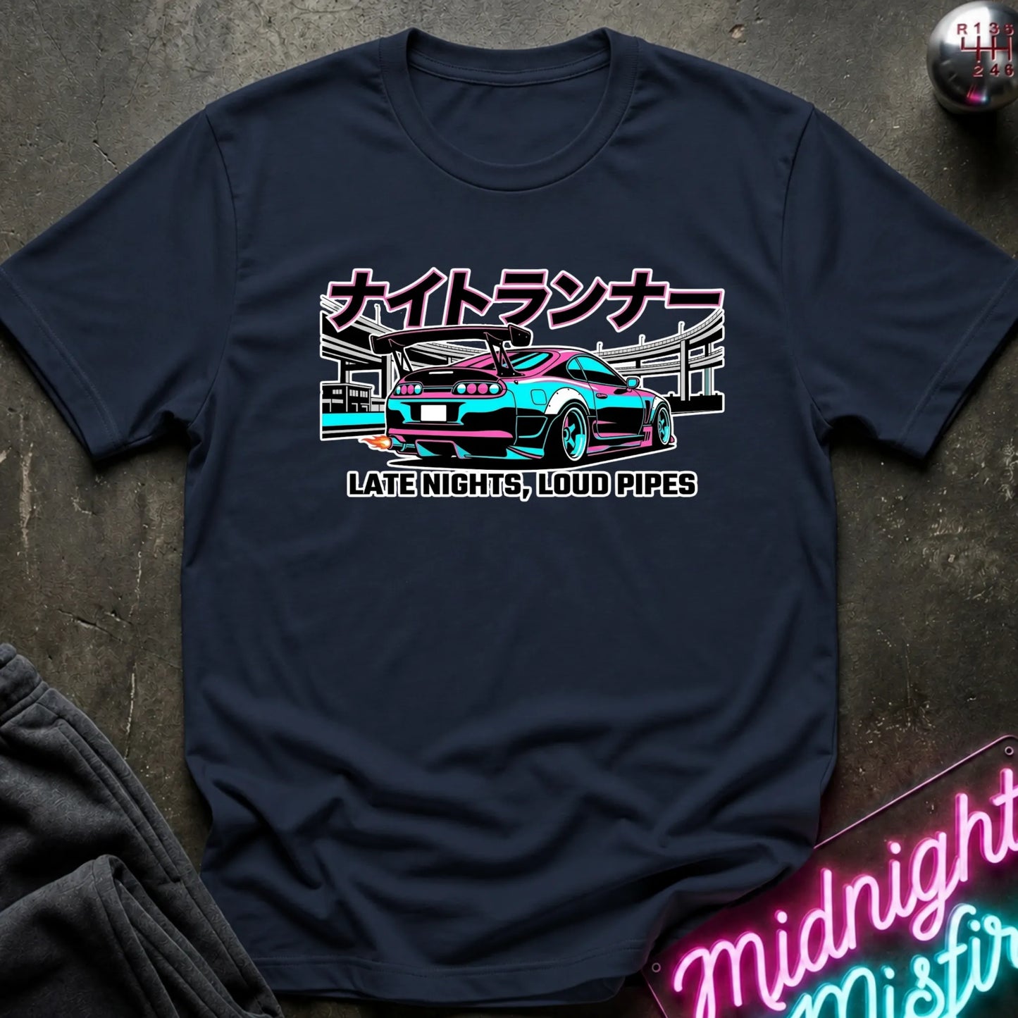 A navy blue graphic t-shirt laid flat on a dark textured surface. The shirt features a colorful retro-style illustration of a sports car with Japanese text above and the phrase 'LATE NIGHTS, LOUD PIPES' below.