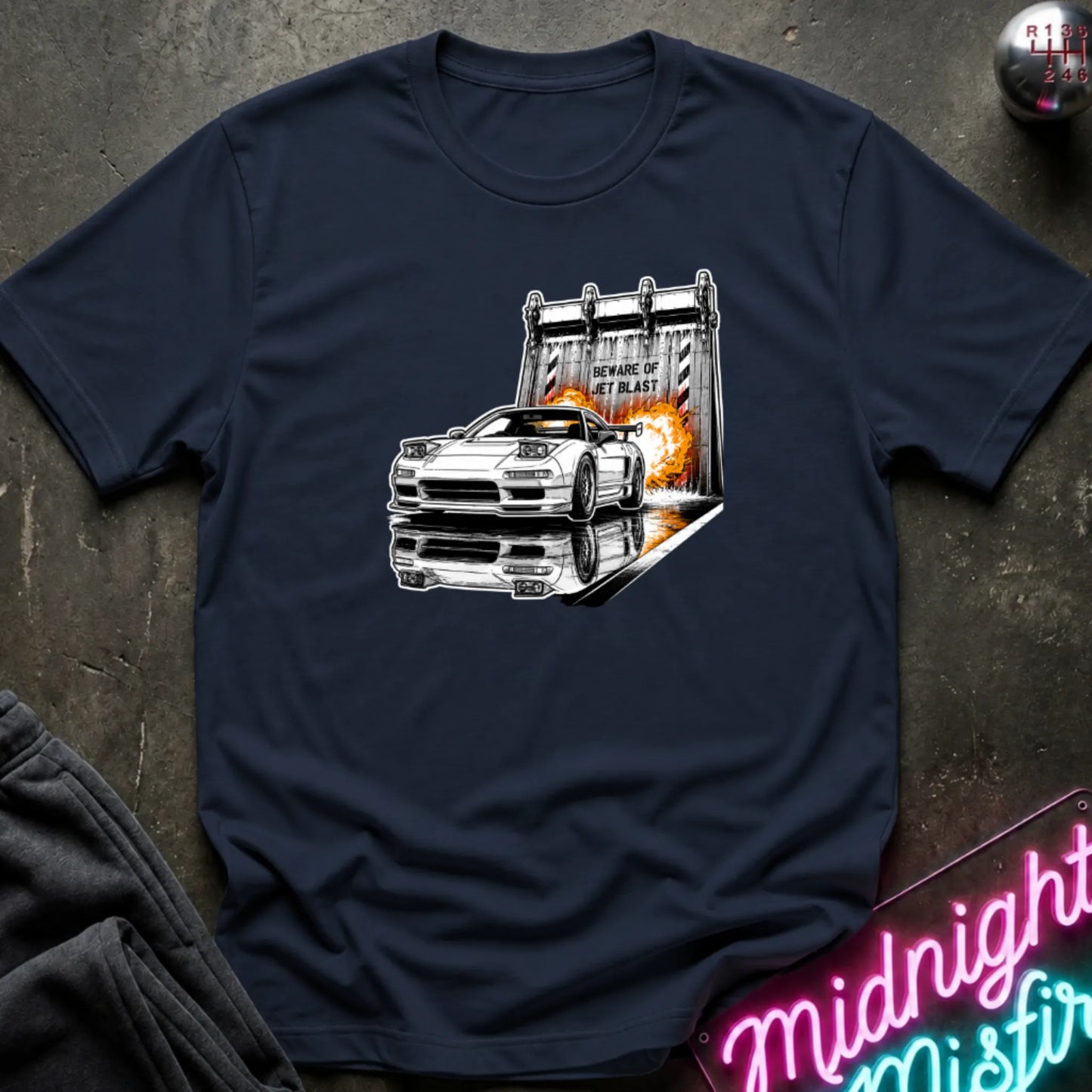 A navy short-sleeve t-shirt laid flat on a dark textured surface, featuring a graphic print of a white sports car in front of a blast off area.