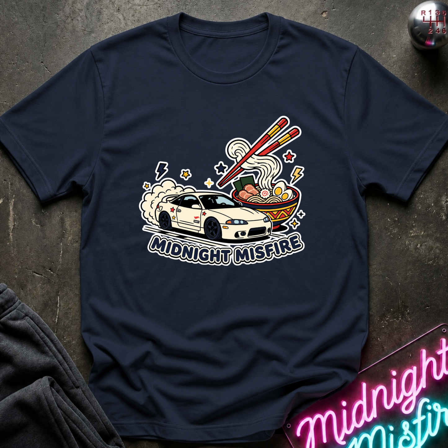 A navy blue graphic t-shirt laid flat on a dark textured surface. The shirt features a colorful graphic of a white sports car driving through steam and smoke with chopsticks over ramen, and the text 'MIDNIGHT MISFIRE' printed below.