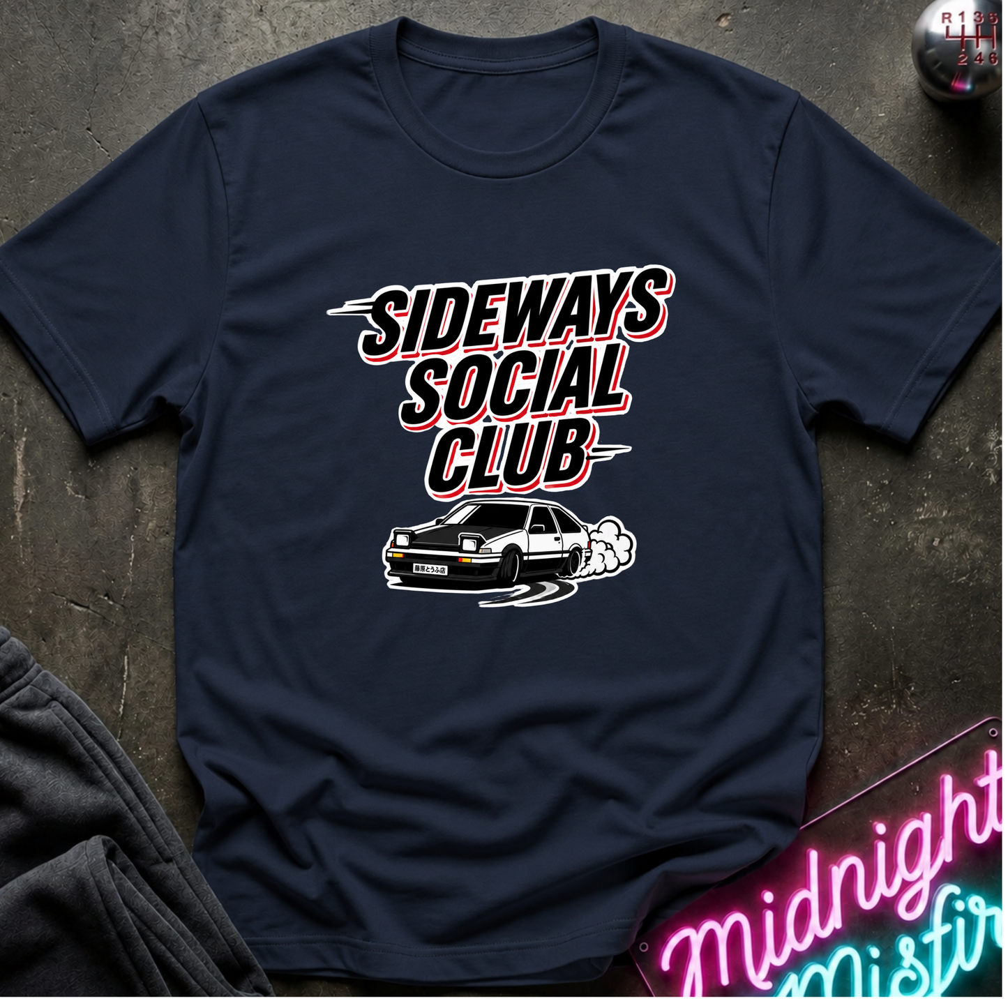 A navy blue graphic t-shirt laid flat on a dark textured surface. The shirt features a graphic print with the text 'SIDEWAYS SOCIAL CLUB' in a stylized white and red font, above an illustration of a white car drifting with smoke.