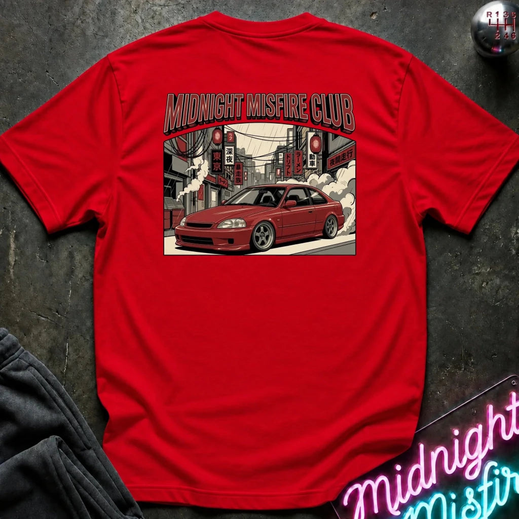 A bright red short-sleeve t-shirt laid flat on a dark textured surface, showing the back graphic with the text 'MIDNIGHT MISFIRE CLUB' above an illustration of a red car in a city street. 