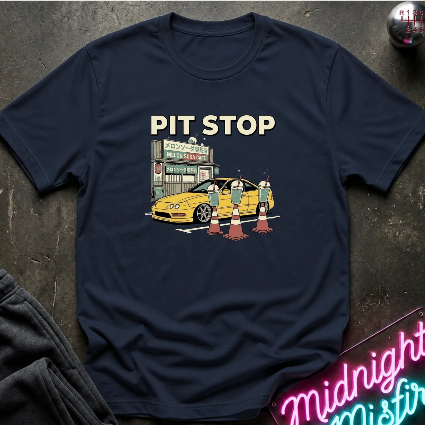 A navy blue graphic t-shirt laid flat on a dark textured surface. The graphic features the text 'PIT STOP' above a yellow car stopped at a gas station with traffic cones in front.