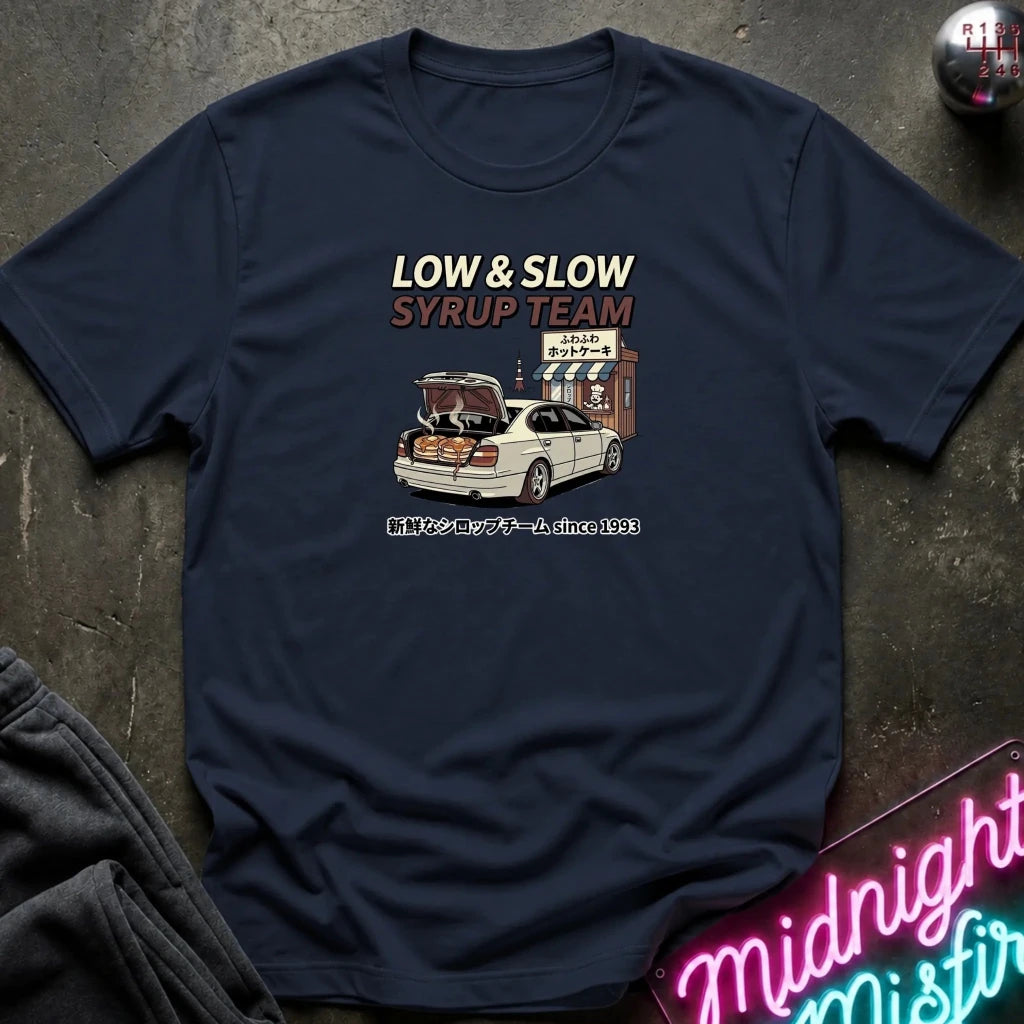 A navy blue crewneck t-shirt laid flat on a dark textured surface. The shirt features a graphic print with the text 'LOW & SLOW SYRUP TEAM' in cream and brown, an illustration of a white car with its trunk open, and Japanese text below.