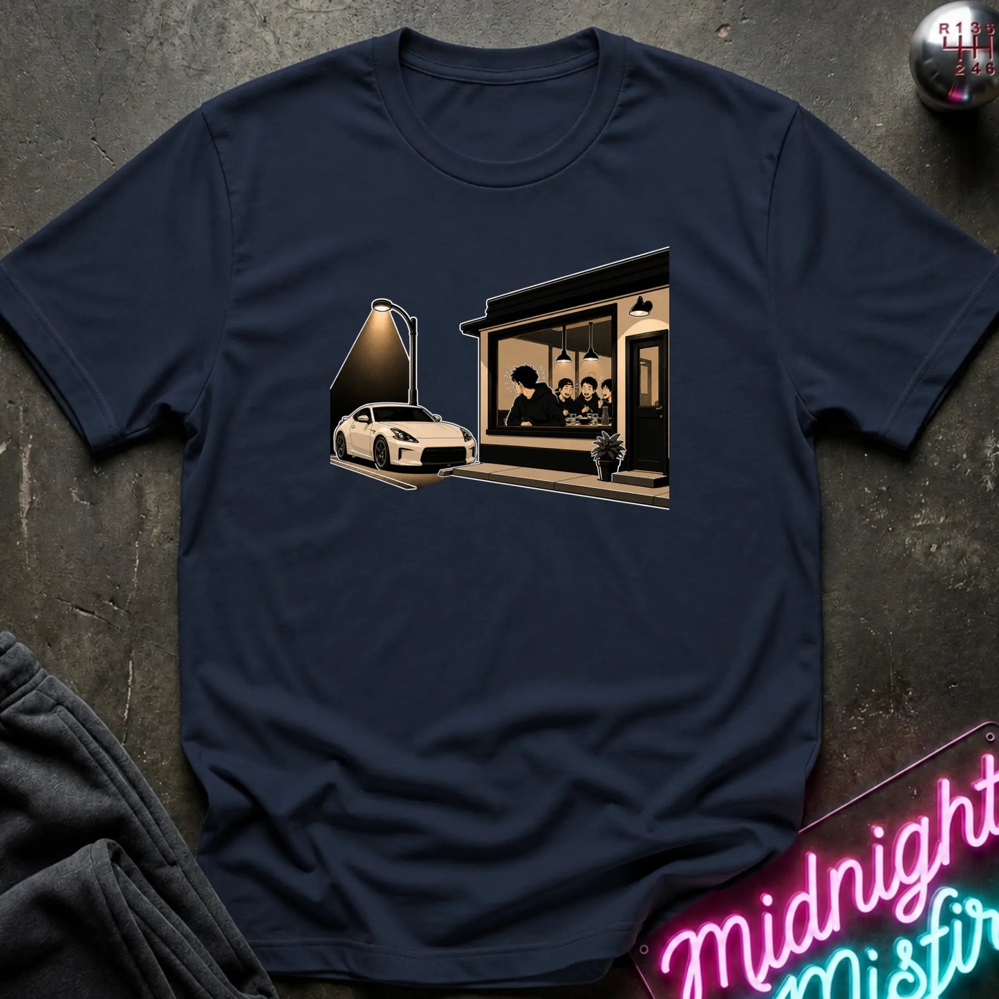 A navy blue short-sleeve t-shirt with a sepia-toned graphic print of a sports car parked outside a cafe, laid flat on a dark textured surface with a neon sign and folded sweatpants nearby.