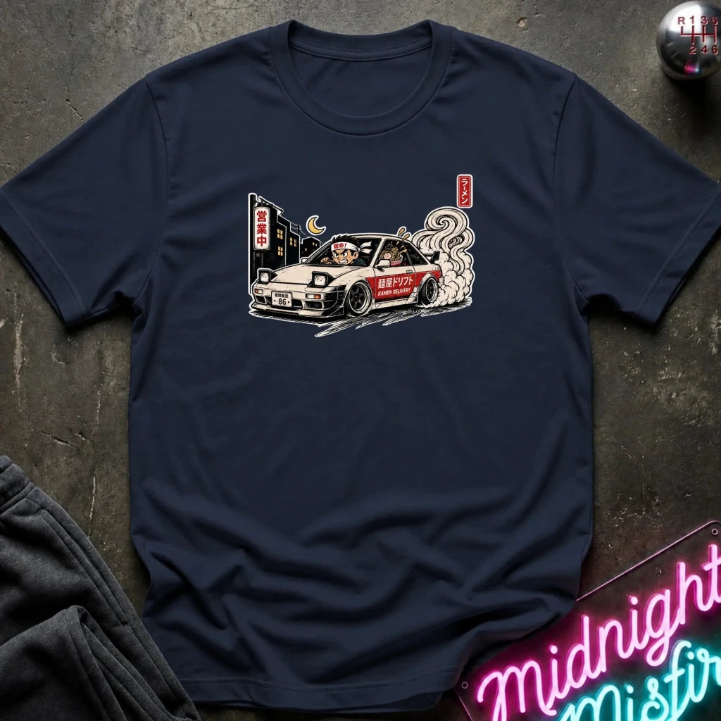 A navy blue short-sleeve t-shirt with a graphic print of a white sports car in a Japanese street scene, laid flat on a dark textured surface with a neon sign and dark pants in the background.