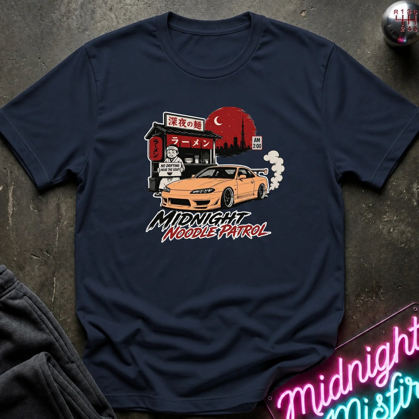 A navy blue short-sleeve t-shirt laid flat on a dark textured surface, featuring a colorful graphic print of an orange sports car in front of a ramen shop at night with the text 'Midnight Noodle Patrol'.