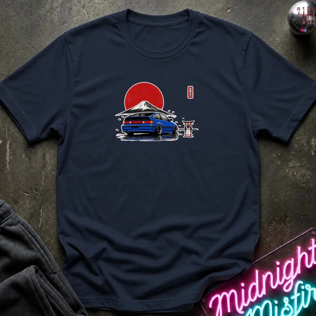 A navy blue short-sleeve t-shirt laid flat on a dark textured surface, featuring a graphic print of a blue sports car in front of a red sun and mountain with a small red vertical text element. 