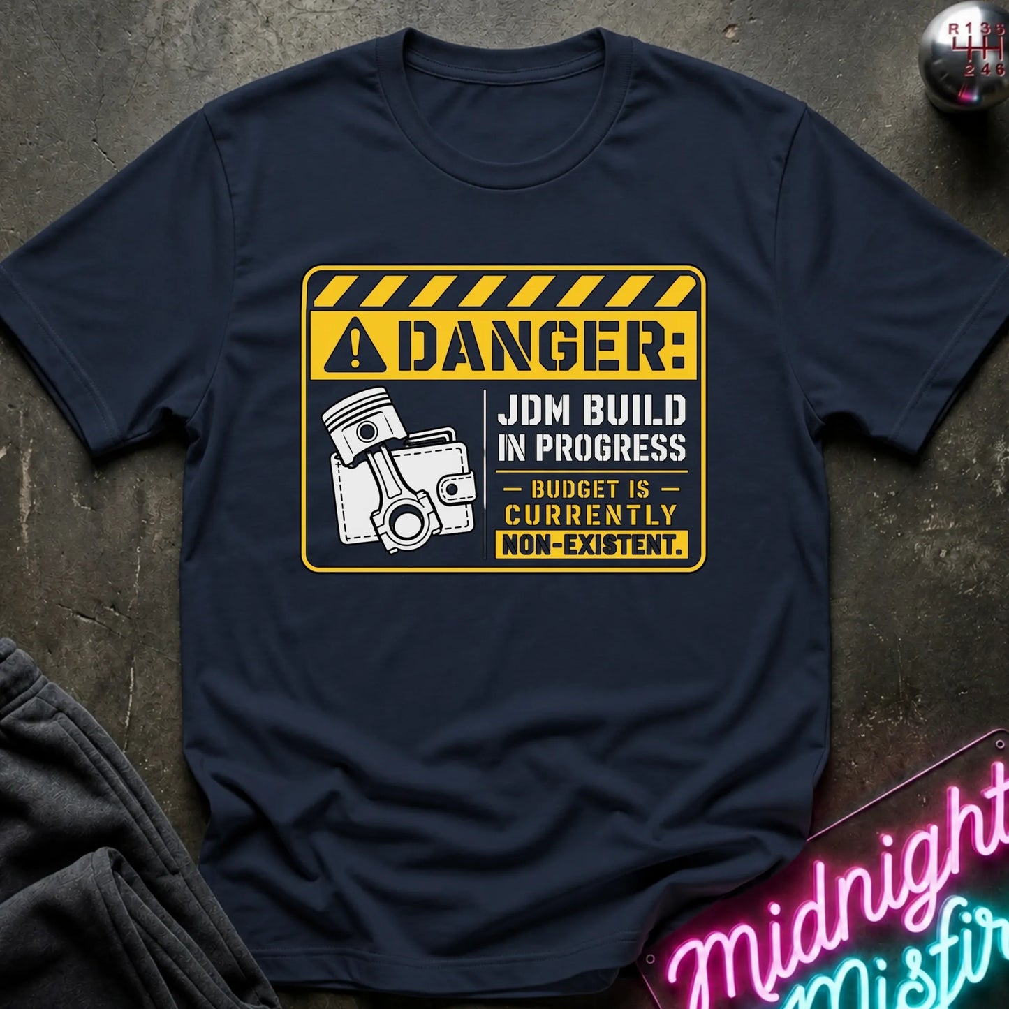 A navy blue short-sleeve graphic T-shirt laid flat on a dark textured surface. The shirt features a yellow and white warning-sign style print with the text 'DANGER: JDM BUILD IN PROGRESS - BUDGET IS - CURRENTLY NON-EXISTENT'.
