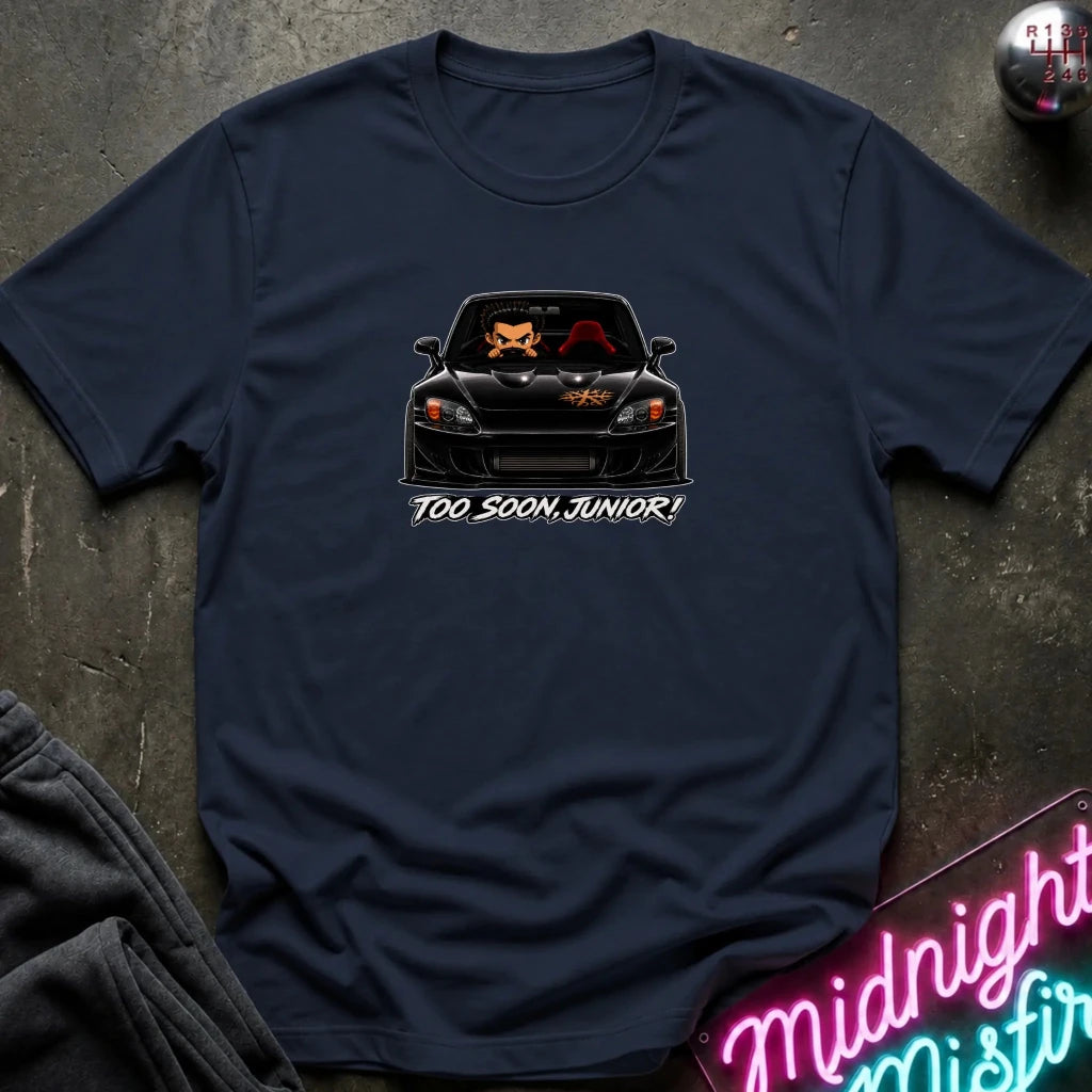 A navy short-sleeve t-shirt laid flat on a dark textured surface, featuring a graphic of a black sports car with orange and red details and the text 'TOO SOON, JUNIOR!' printed below it.