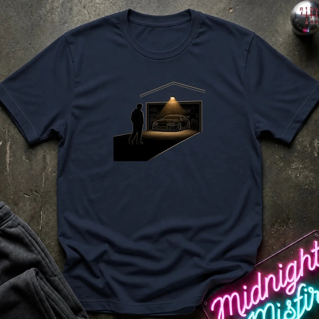 A navy blue crewneck t-shirt laid flat on a dark textured surface, featuring a graphic print of a silhouette of a man looking at a sports car inside a garage with a spotlight.