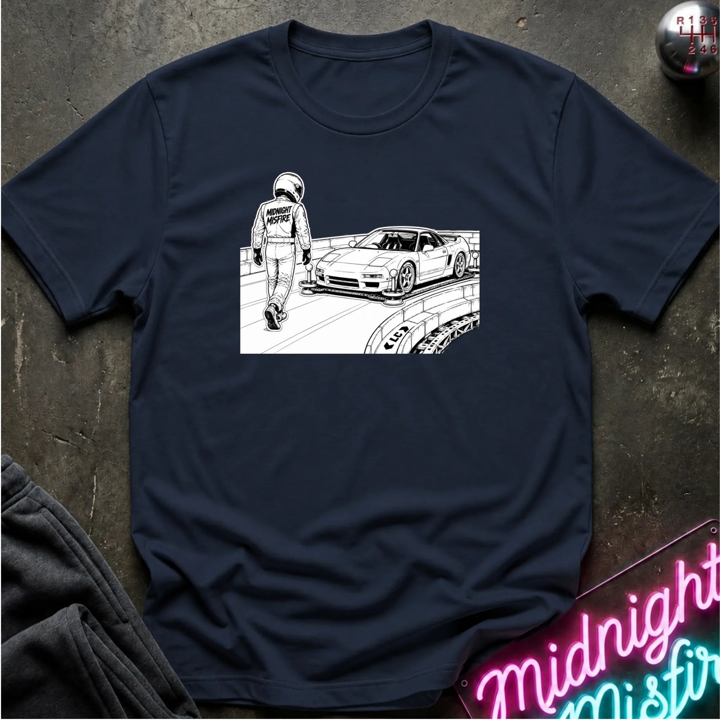 A navy blue short-sleeve t-shirt with a black and white graphic print of a race car and a person in a racing suit, laid flat on a dark textured surface.