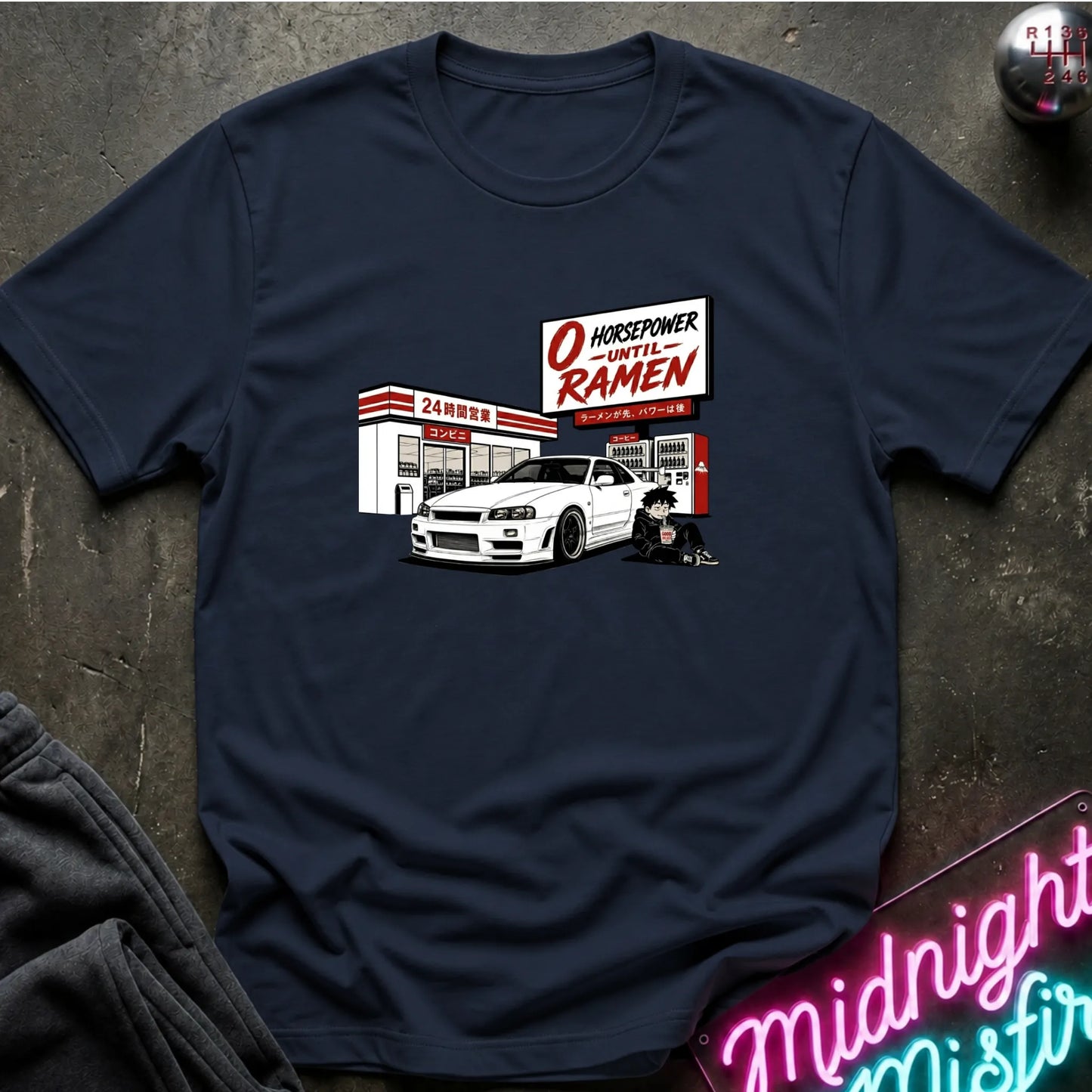 A navy blue graphic t-shirt laid flat on a dark textured surface. The graphic features a white sports car parked in front of a store with a sign reading 'O HOURSEPOWER UNTIL RAMEN'.