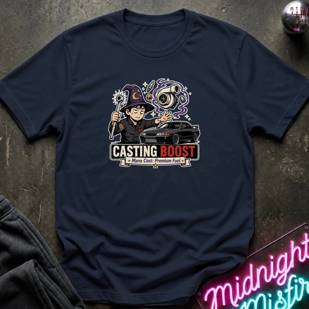 A navy blue graphic t-shirt laid flat on a dark textured surface. The graphic features a cartoon character holding a pointer and a key, next to a black car, with the text 'CASTING BOOST' and 'Many Costs - Premium Fuel'.