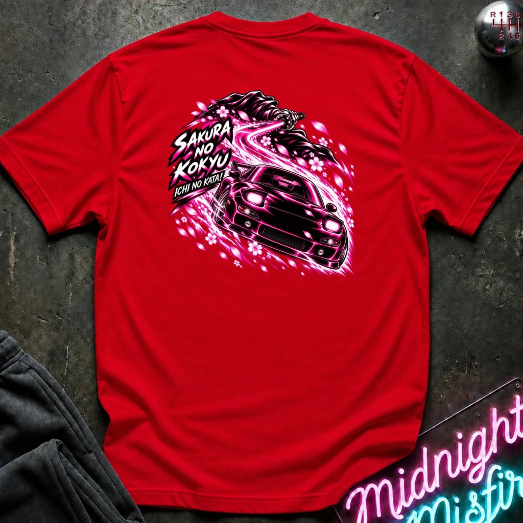 A red short-sleeve t-shirt laid flat on a dark textured surface, showing the back graphic with a car and the text "ichi no kata!"