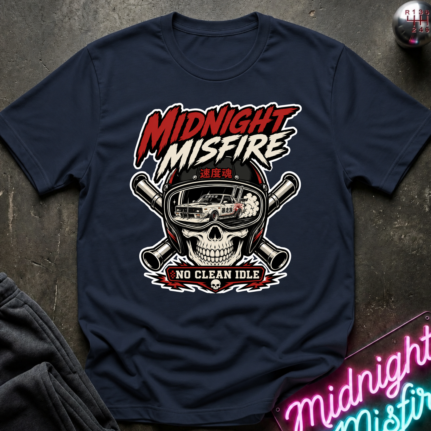 A navy blue short-sleeve t-shirt with a graphic print featuring a skull wearing a helmet, crossed guns, and the text 'MIDNIGHT MISFIRE' and 'NO CLEAN IDLE'. The shirt is laid flat on a dark textured surface with a neon sign and dark fabric visible in the corners.