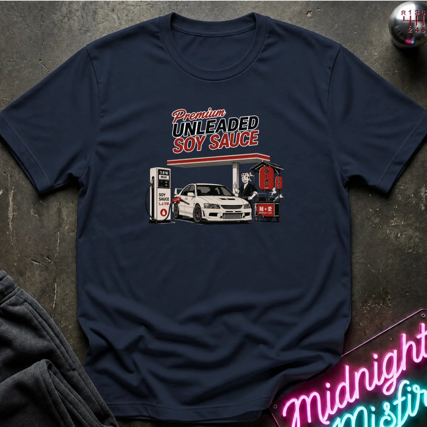 A navy short-sleeve graphic t-shirt laid flat on a dark textured surface. The graphic features a vintage-style illustration of a gas station attendant filling up a white car with soy sauce, with the text 'Premium UNLEADED SOY SAUCE' above it.