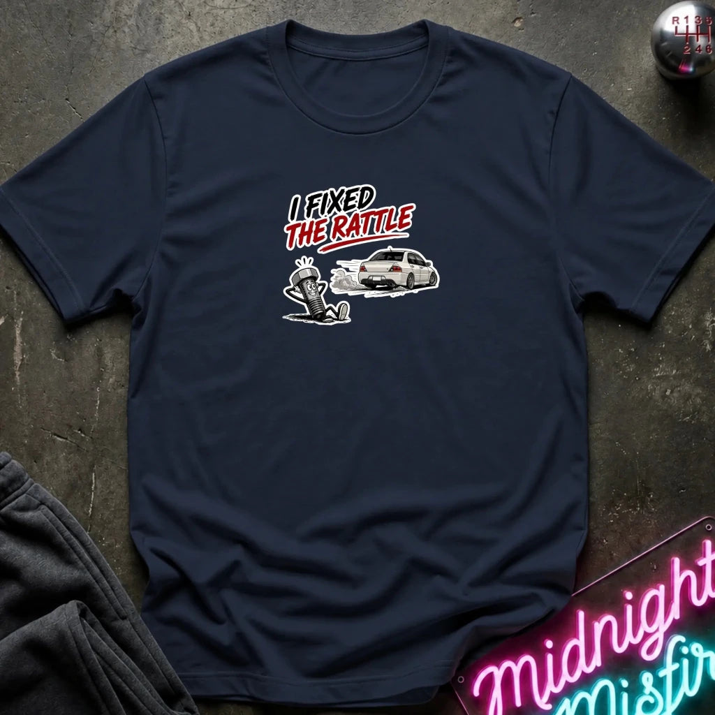 A navy blue short-sleeve t-shirt with a graphic print on the chest that reads 'I FIXED THE RATTLE' above a white car and a hand standing on a curb. The shirt is laid flat on a dark textured surface with a neon sign and folded sweatpants partially visible.