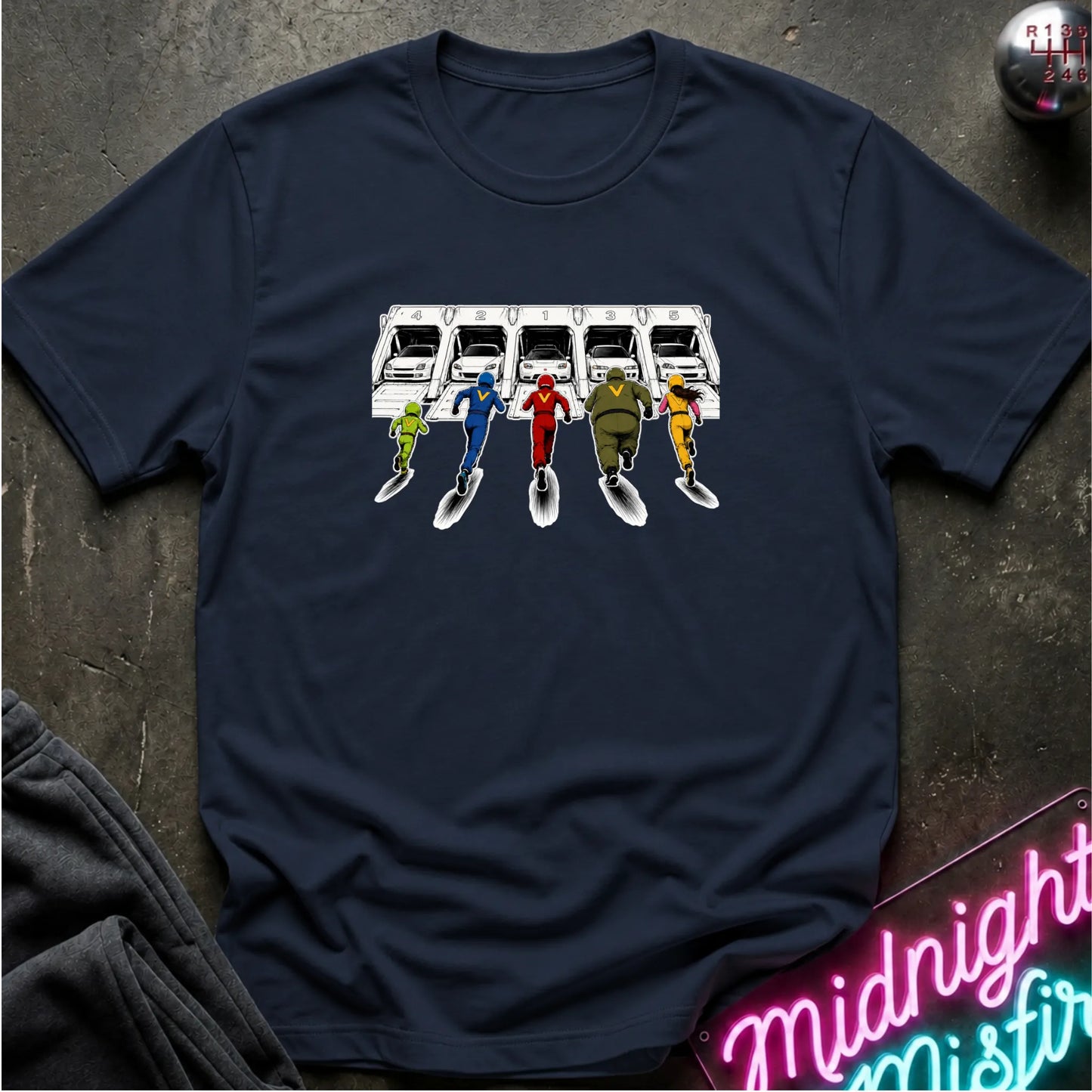 A navy blue short-sleeve graphic T-shirt laid flat on a dark textured surface. The shirt features a printed illustration of a white sports cars in 5 different launch  pods.