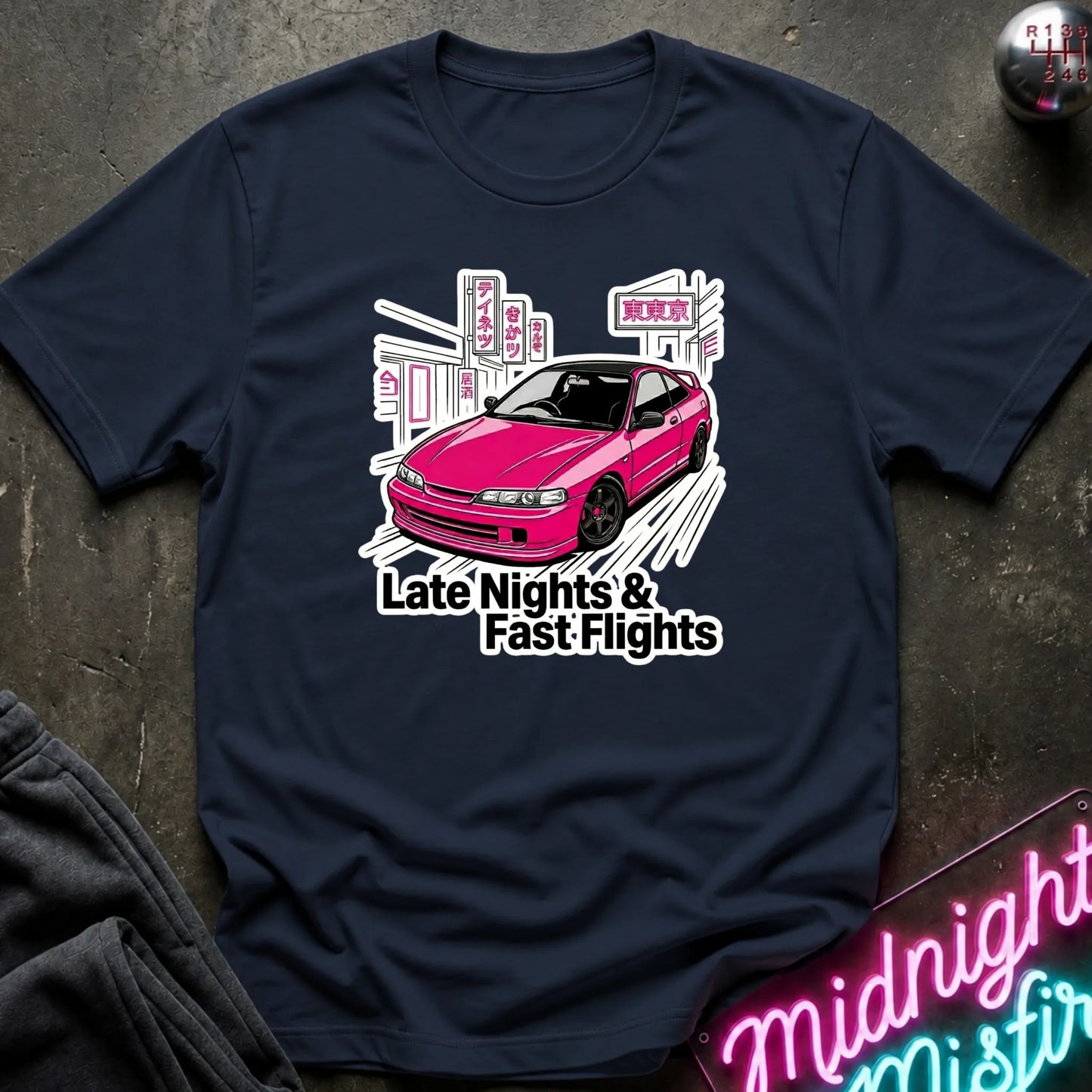 A navy blue graphic t-shirt laid flat on a dark textured surface. The shirt features a graphic of a pink car in front of a white cityscape with the text 'Late Nights & Fast Flights'.