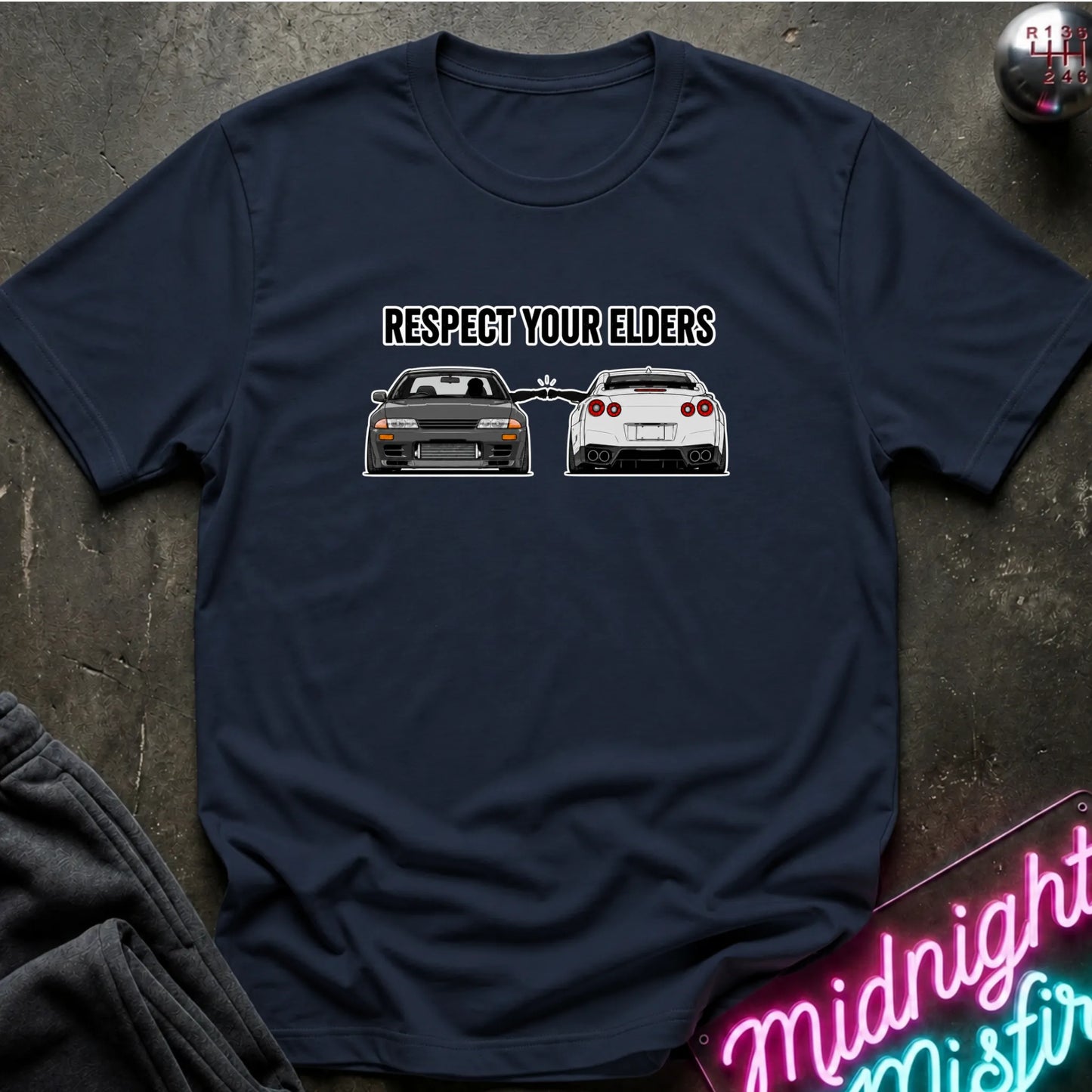 A navy blue short-sleeve t-shirt laid flat on a dark textured surface. The shirt features a graphic print of a black car in front of a white car with the text 'RESPECT YOUR ELDERS' above them.