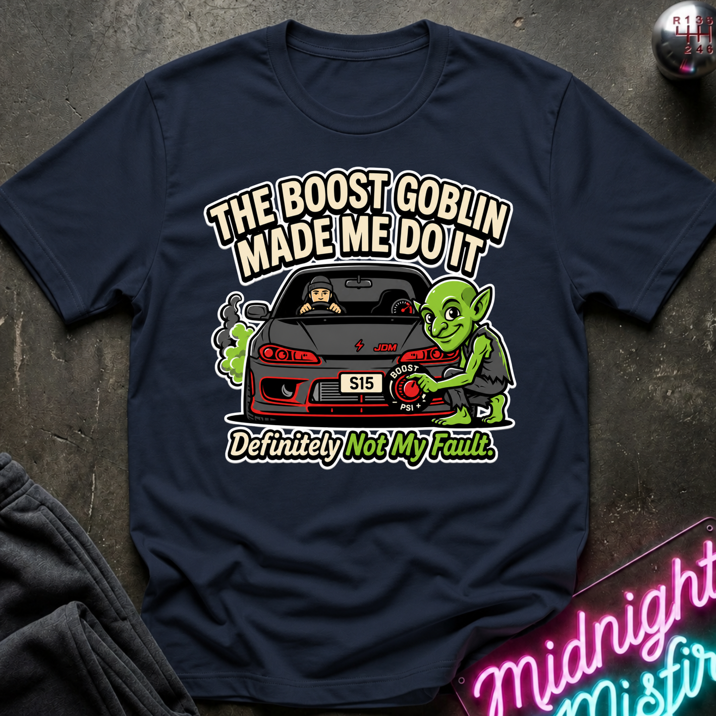 A navy blue graphic t-shirt laid flat on a dark textured surface. The shirt features a cartoon illustration of a green goblin character next to a car, with the text 'THE BOOST GOBLIN MADE ME DO IT' and 'Definitely Not My Fault.