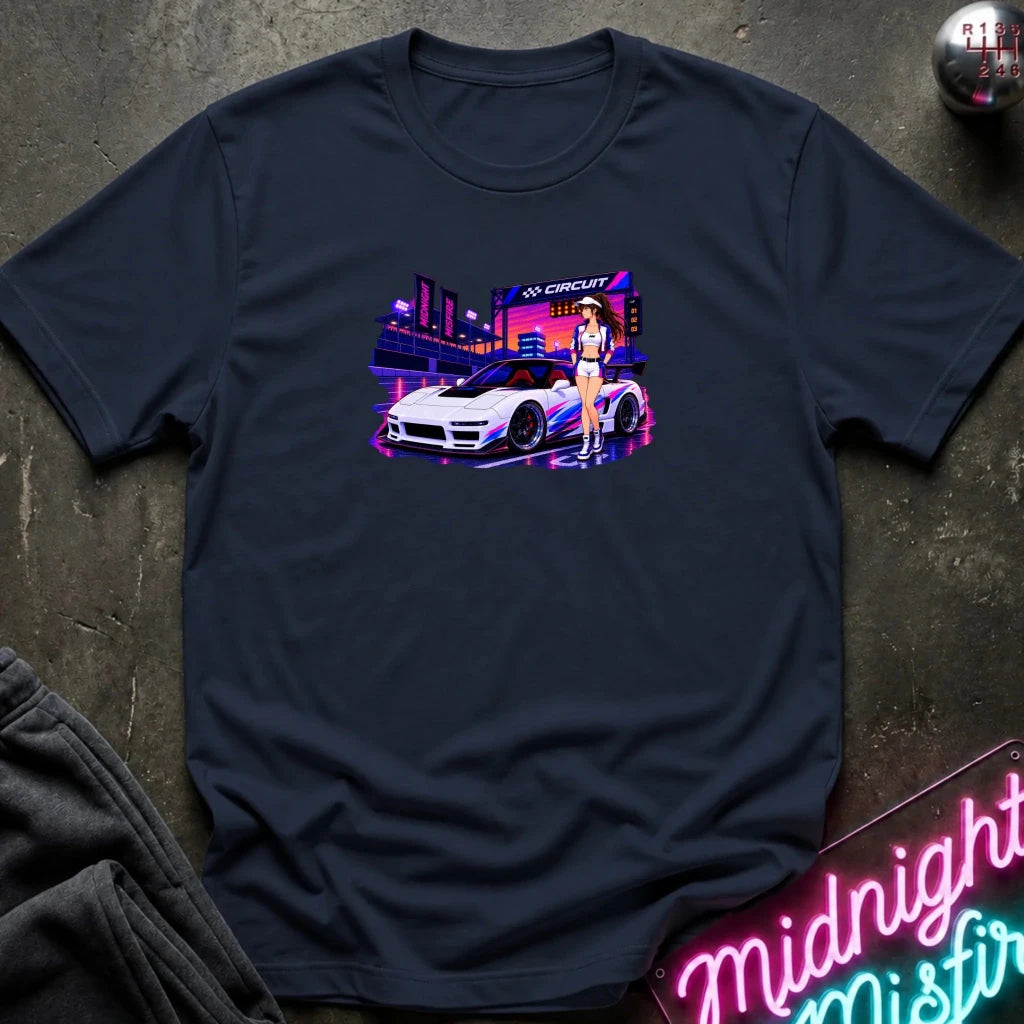 A navy blue short-sleeve t-shirt with a colorful graphic print of a white sports car and a woman in a neon cityscape, laid flat on a dark textured surface with a neon sign and sweatpants nearby.