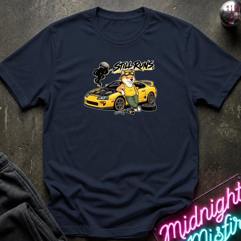 A navy blue graphic t-shirt laid flat on a dark textured surface. The graphic features a cartoon dog in a mechanic hat working on a yellow sports car with the text 'STILL RUNS' above it.
