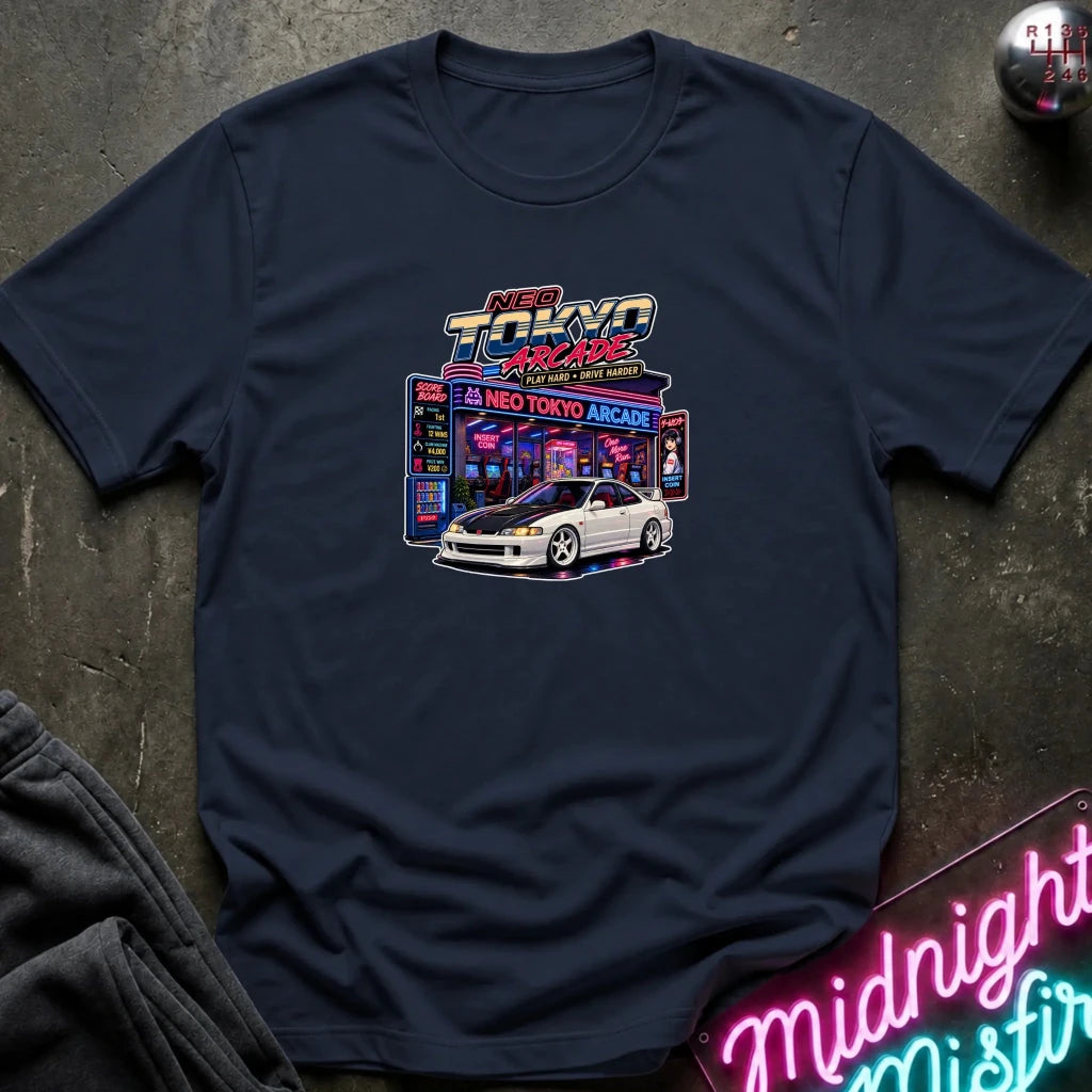 A navy short-sleeve crewneck t-shirt featuring a 'Neo Tokyo Arcade' graphic with a white sports car, laid flat on a dark textured surface with a neon sign and sweatpants as props.