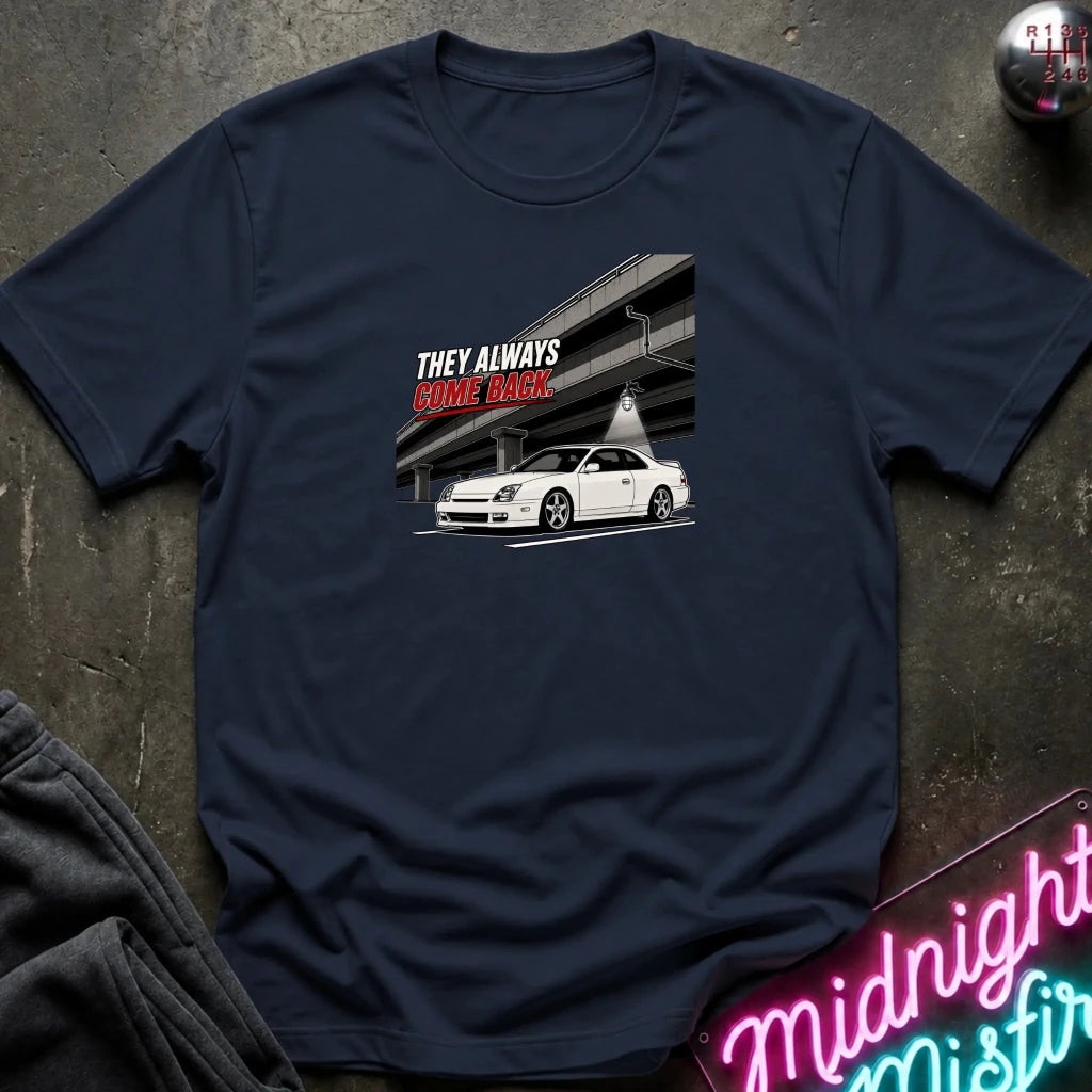 A navy blue short-sleeve t-shirt laid flat on a dark textured surface. The shirt features a graphic print of a white sports car under an overpass with the text 'THEY ALWAYS RACE BACK.