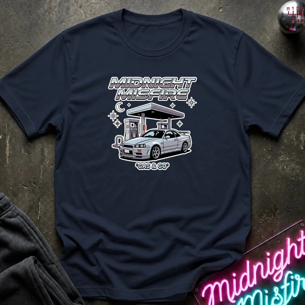 A navy blue graphic short-sleeve t-shirt with a graphic print of a silver Nissan S13 'Kits & Co' car at a gas station under the text 'MIDNIGHT MISFIRE' and 'gas & GO', laid flat on a dark textured surface.