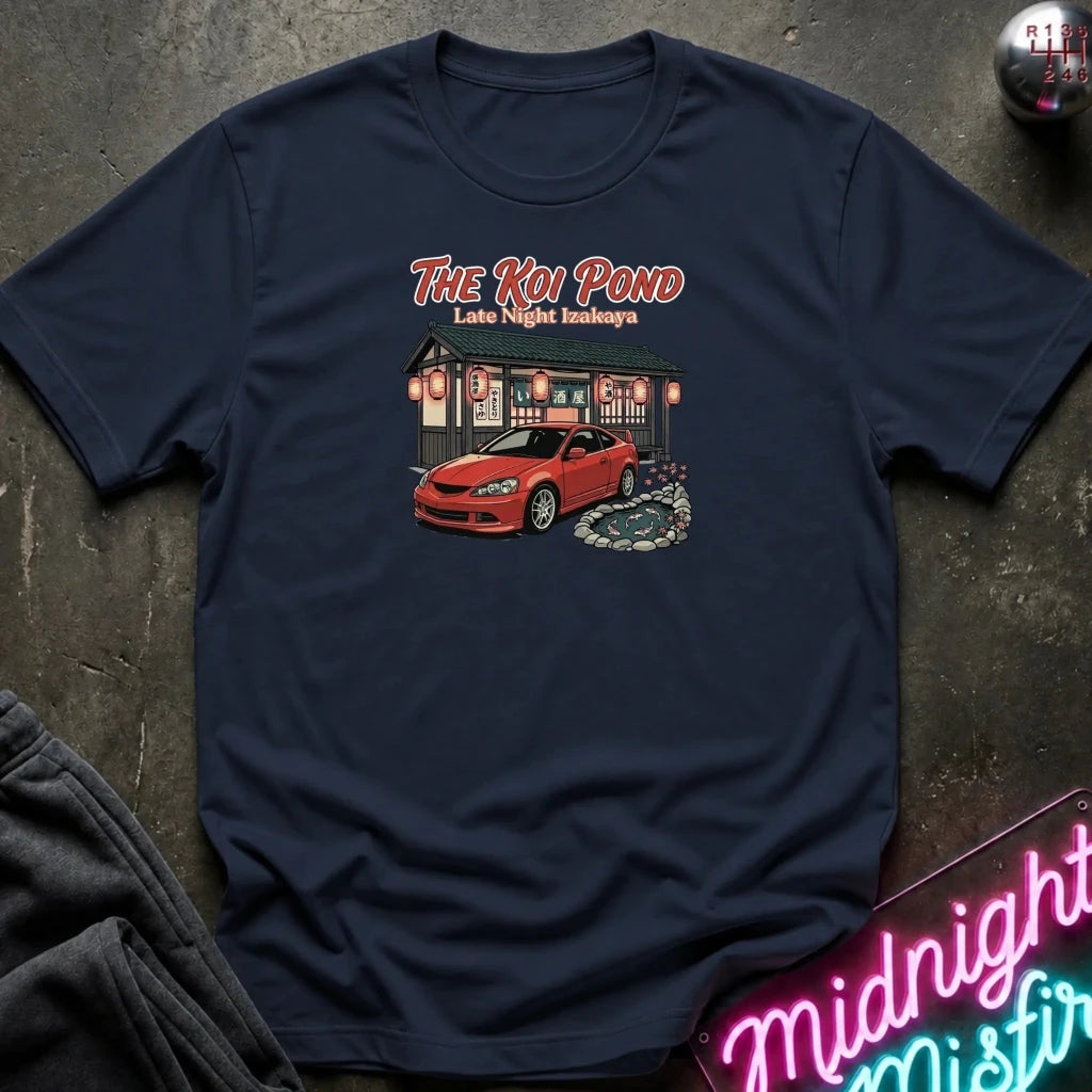 A navy blue short-sleeve graphic t-shirt laid flat on a dark textured surface. The graphic features the text 'THE KOI POND Late Night Hamburger' above an illustration of a red sports car parked in front of a restaurant with red lanterns and a koi pond.