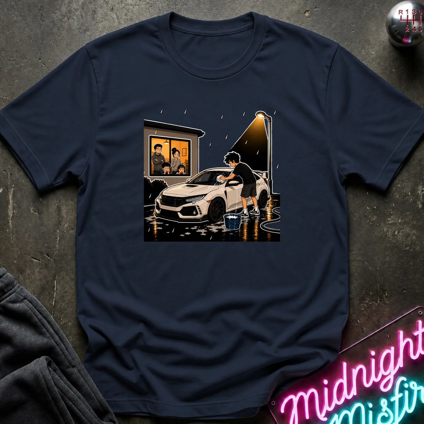 A navy blue short-sleeve t-shirt with a central graphic print of a person washing a white sports car in the rain, styled flat on a dark textured background with a neon sign and clothing prop.