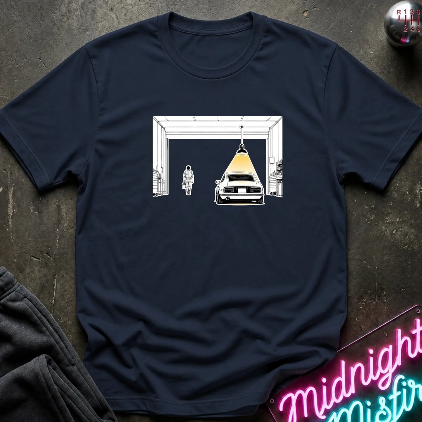A navy blue short-sleeve t-shirt laid flat on a dark textured surface, featuring a graphic print of a car under a hanging lamp inside a garage structure. 
