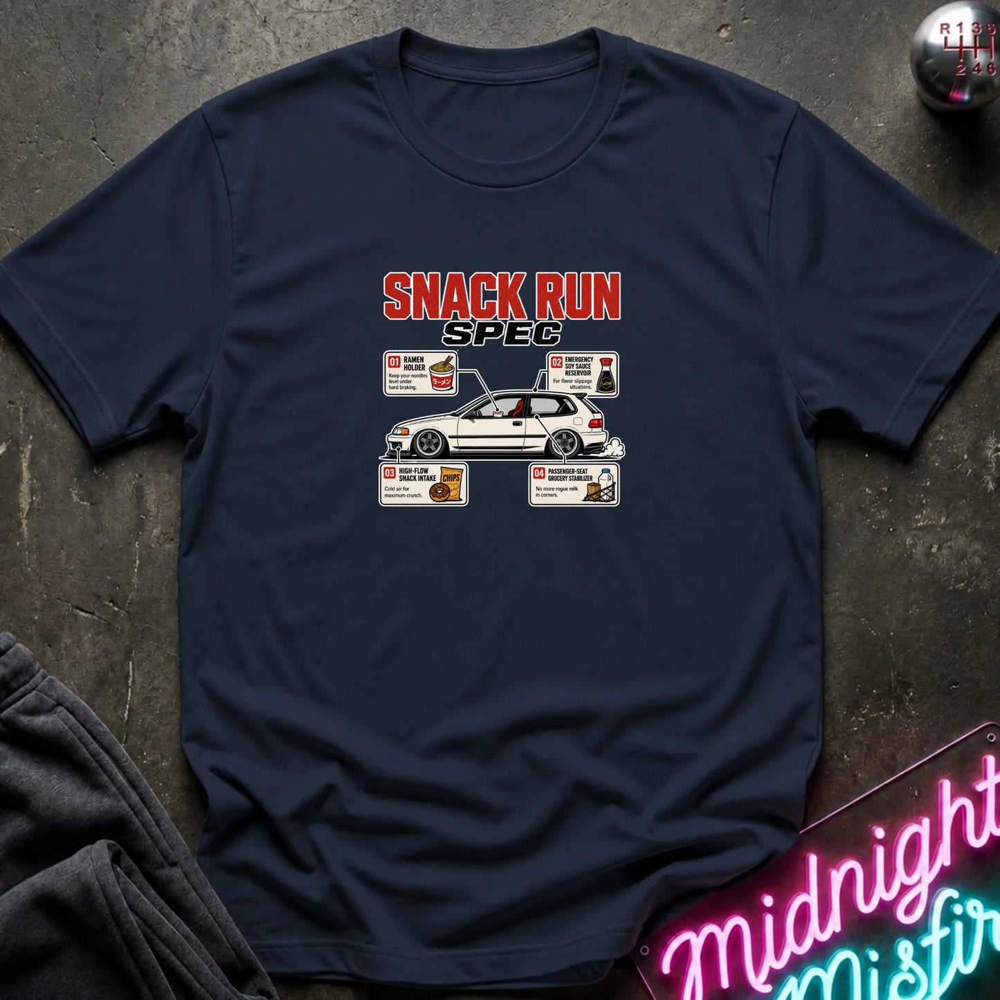 A navy blue graphic t-shirt laid flat on a dark textured surface. The shirt features a 'SNACK RUN SPEC' graphic with a white car and small food boxes.