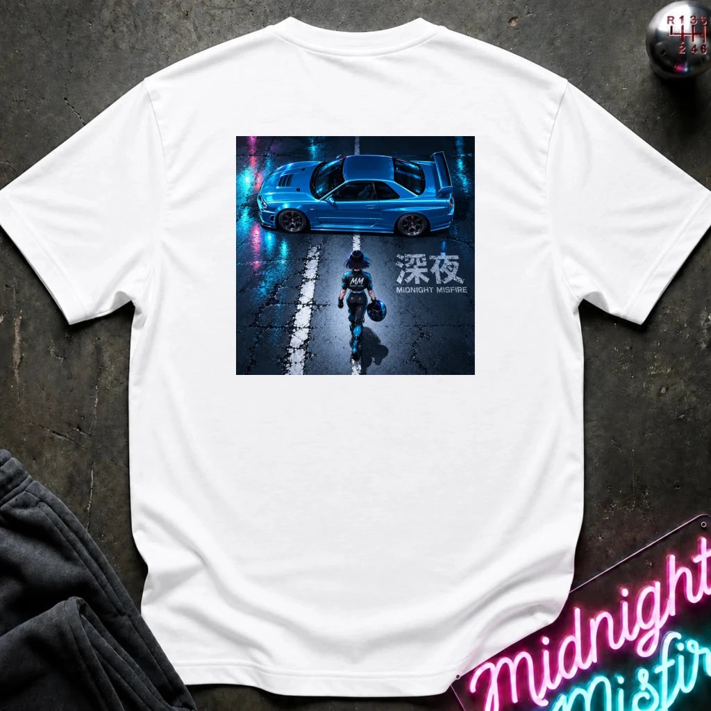 A white short-sleeve t-shirt laid flat on a dark textured surface, featuring a large square graphic print on the back. The graphic shows a blue sports car on a wet street at night with neon reflections and a figure walking toward the camera.
