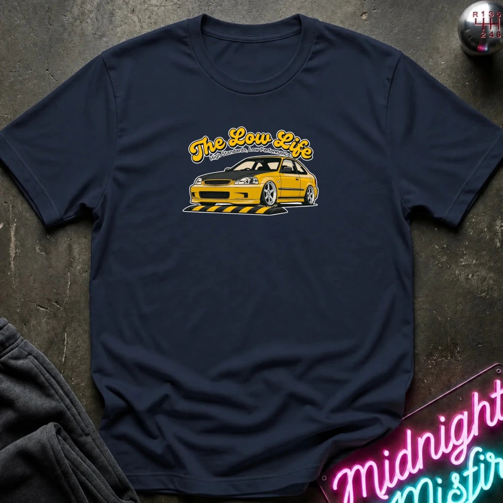 A navy blue graphic t-shirt laid flat on a dark textured surface. The shirt features a yellow and white graphic of a low-profile station wagon with the text 'The Low Life' in yellow script.