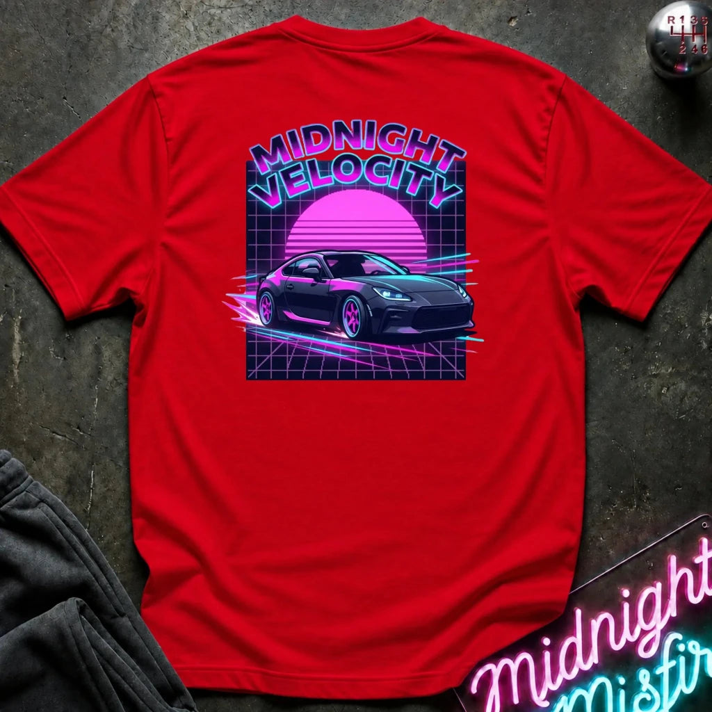 A red short-sleeve t-shirt laid flat on a dark textured surface, featuring a large retro 80s-style graphic on the back with the words 'MIDNIGHT VELOCITY,' a pink sun, a dark sports car, and neon grid lines. Partial black pants and a neon sign are visible around it as styling props.