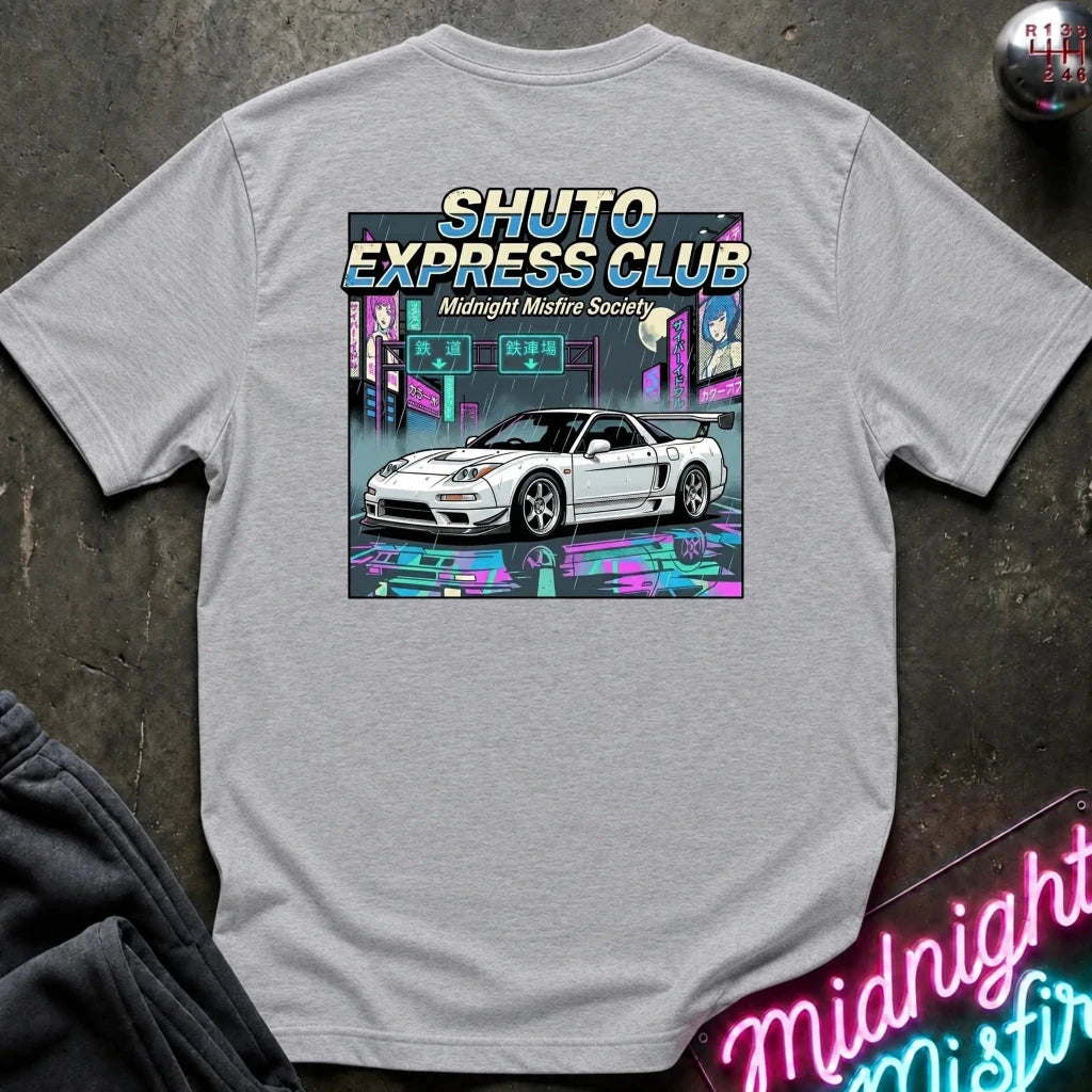A light gray heather crewneck t-shirt featuring a colorful graphic print of a white sports car in a neon cyberpunk city scene with the text 'SHUTO EXPRESS CLUB' and 'Midnight Misfire Society'. The shirt is laid flat on a dark textured surface with a neon sign and dark sweatpants partially visible.